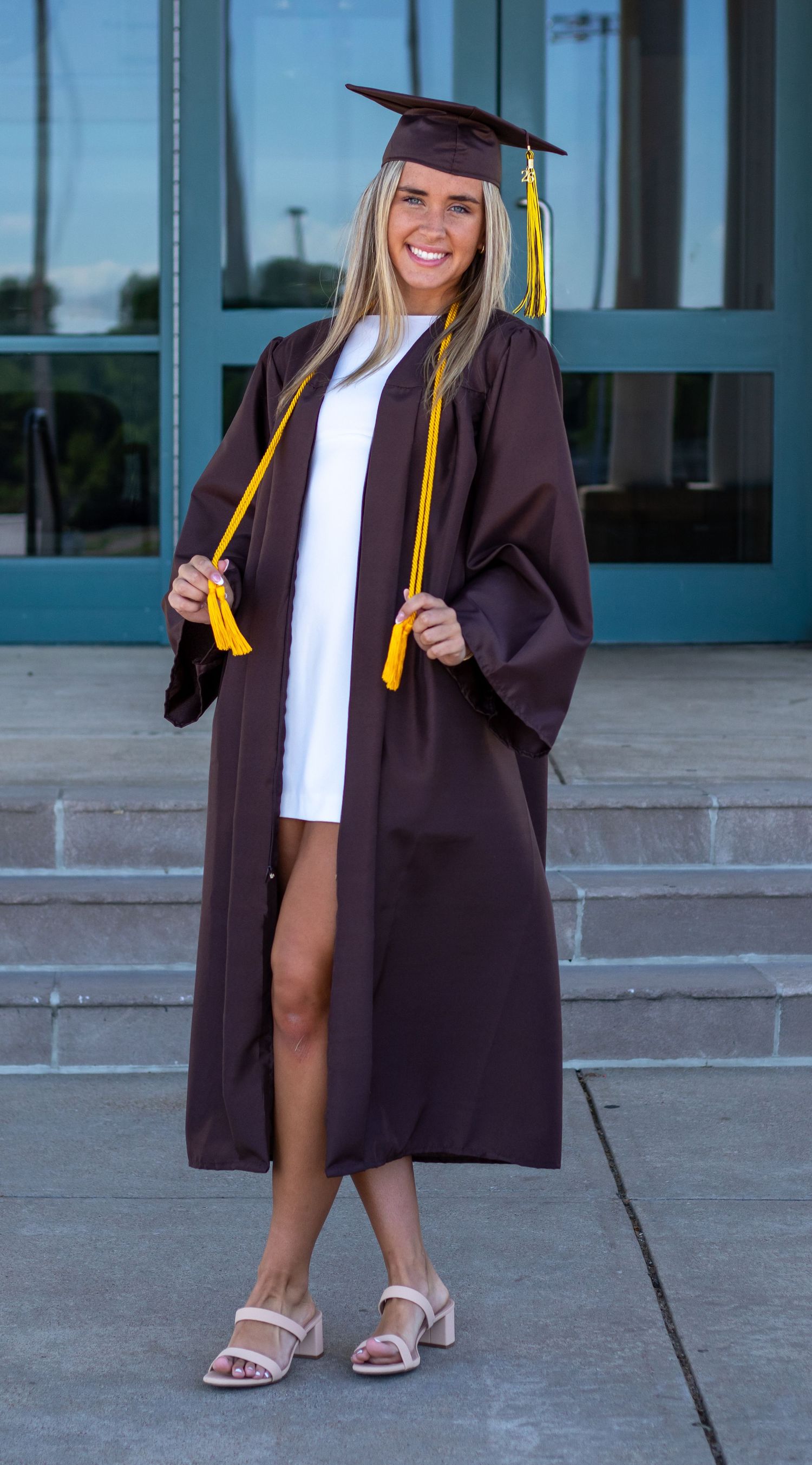 Smiling blonde graduate in brown cap and gown with gold tassels standing on steps.