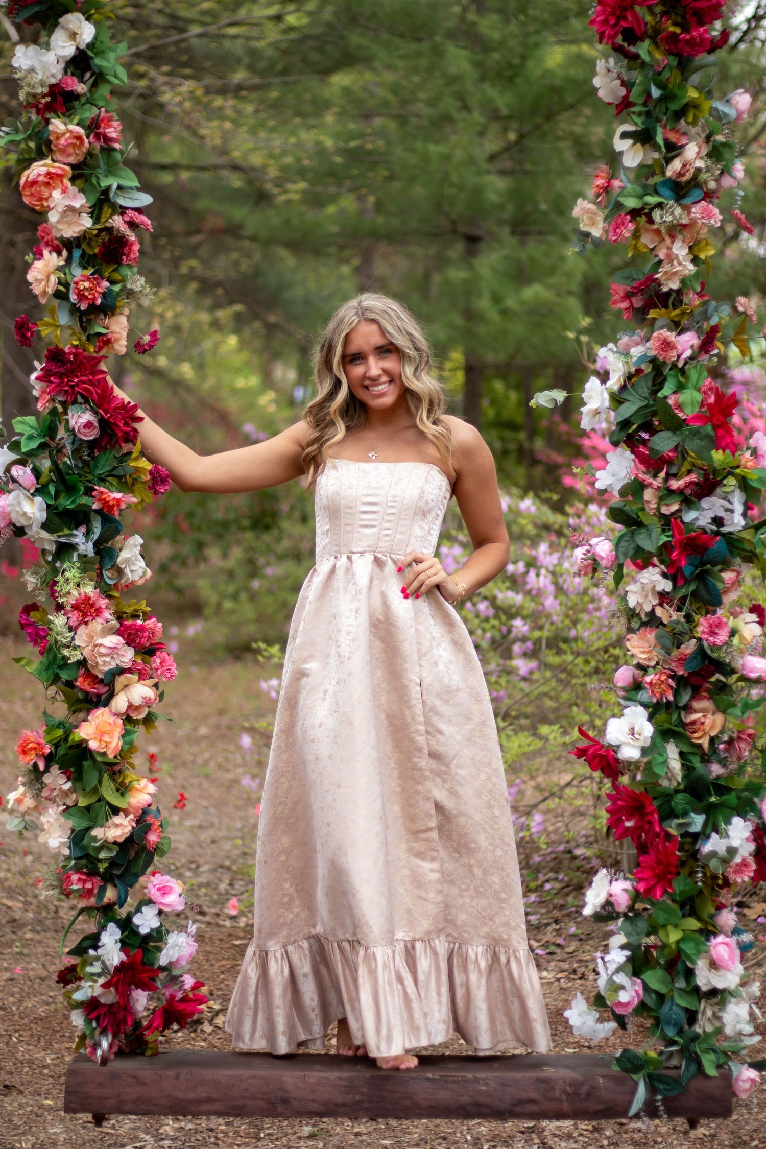 Blonde woman poses elegantly in a strapless blush gown between two floral-covered columns in a garden setting.