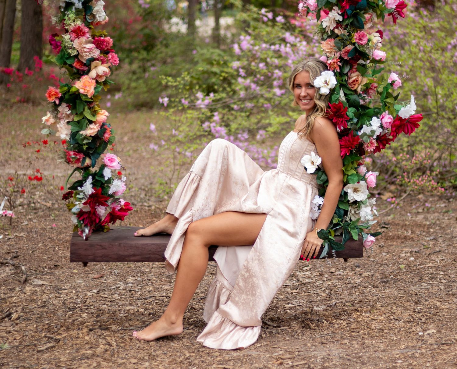 Blonde woman in satin gown sits smiling on a flower-decorated wooden swing in a wooded setting.