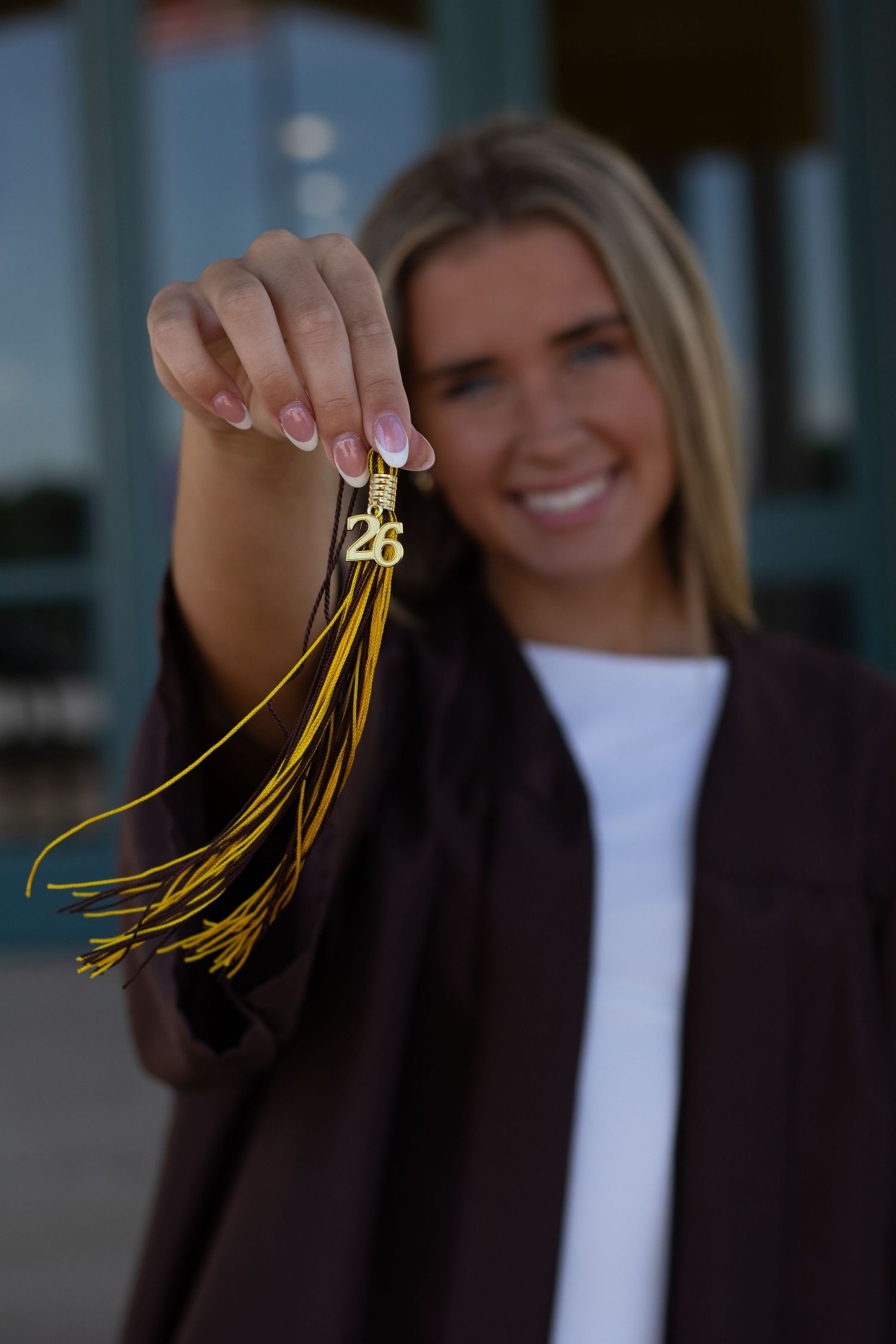 Smiling blonde graduate holding a gold tassel with 26 charm in her graduation gown.