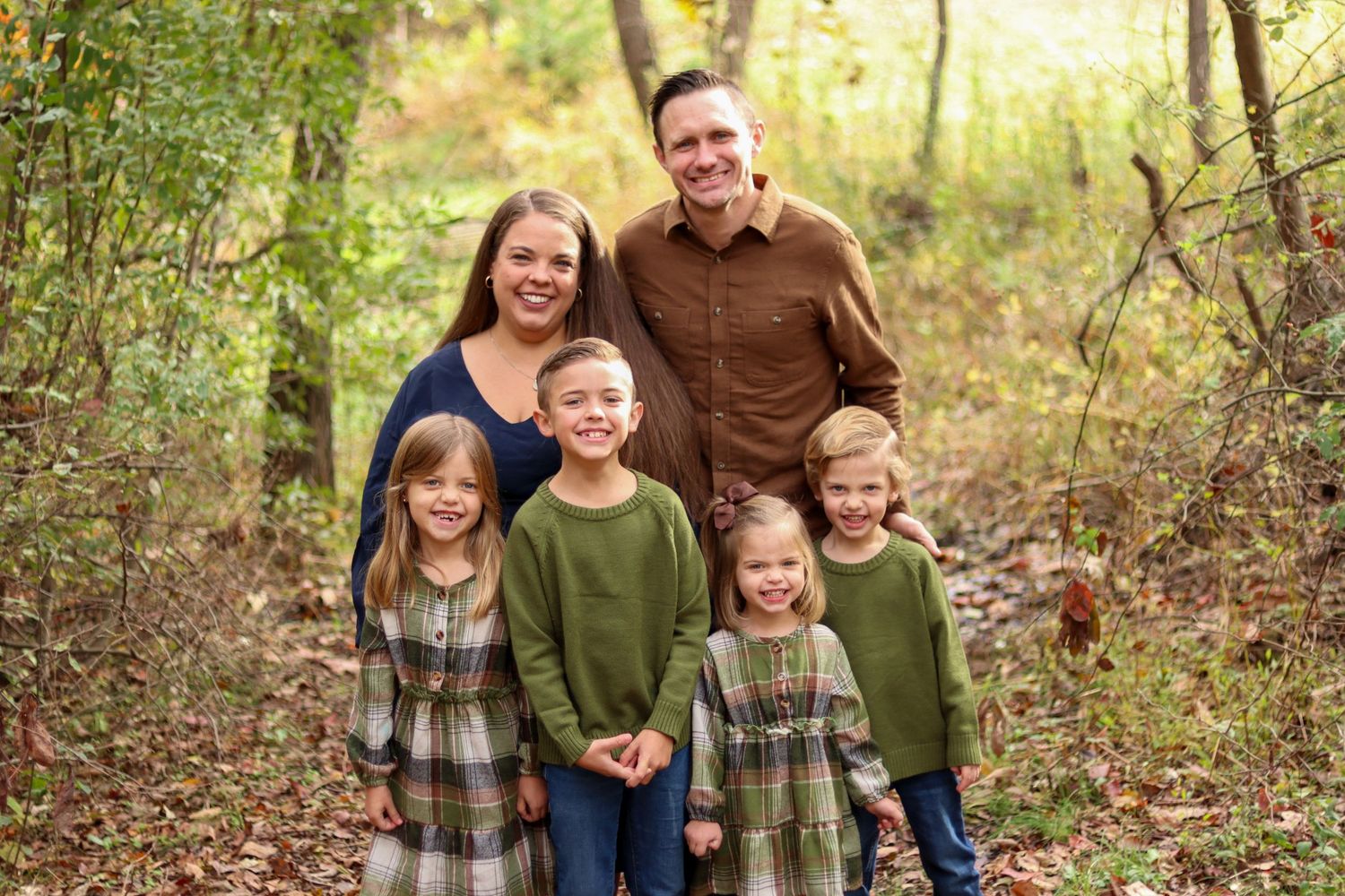 Smiling family of six poses together on wooded trail surrounded by colorful autumn foliage.