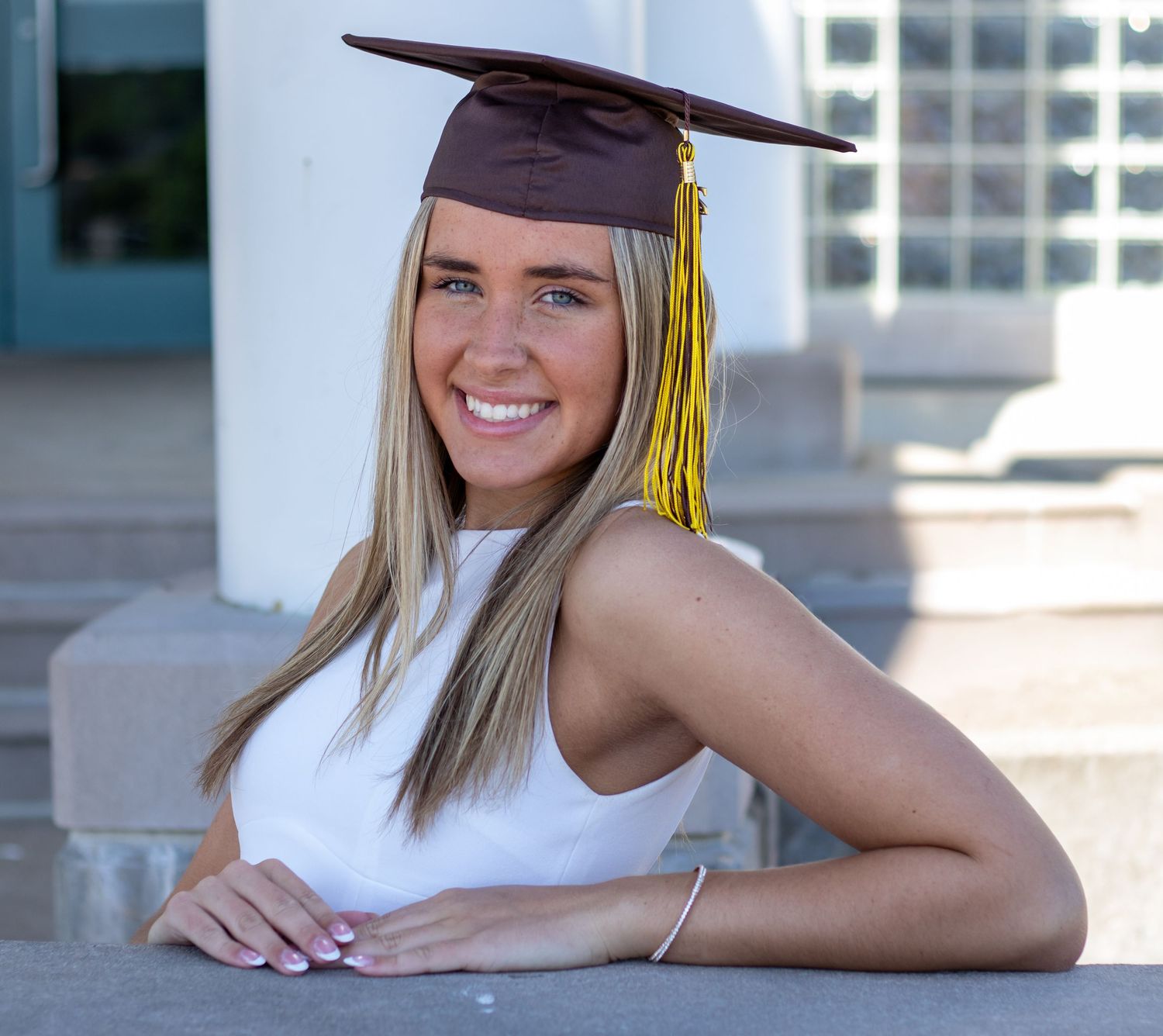 Smiling blonde graduate wearing brown mortarboard cap with gold tassel leaning on stone steps.