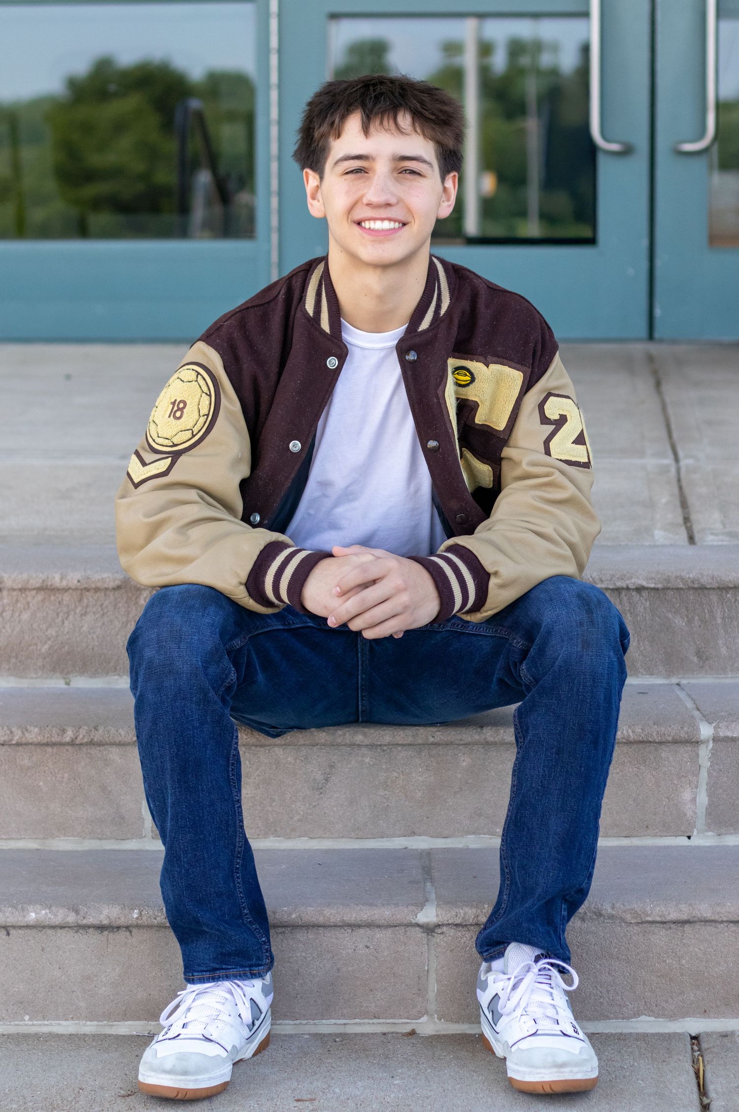 Smiling teen boy in varsity jacket and jeans sitting on school steps for senior photos.