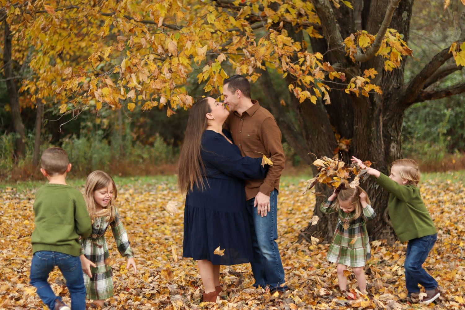 Couple kissing while children play in fallen autumn leaves under tree.