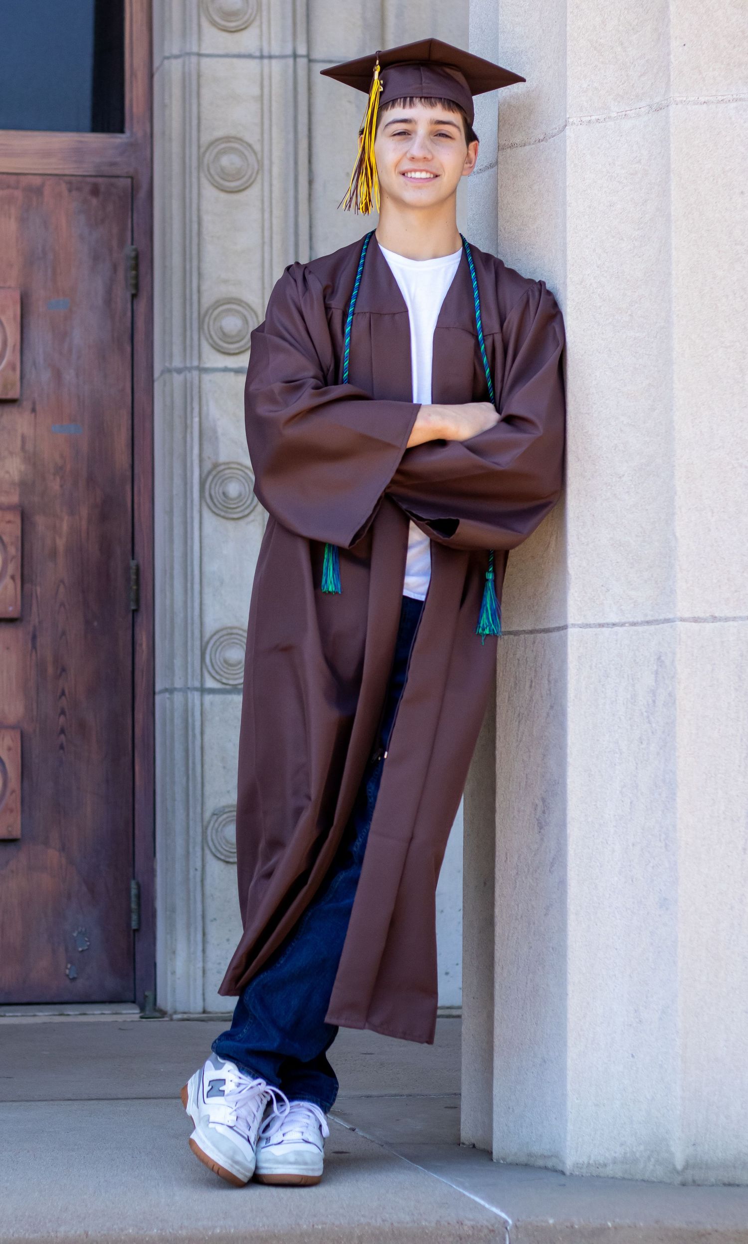 Smiling young male graduate in brown cap and gown poses confidently against stone building column.