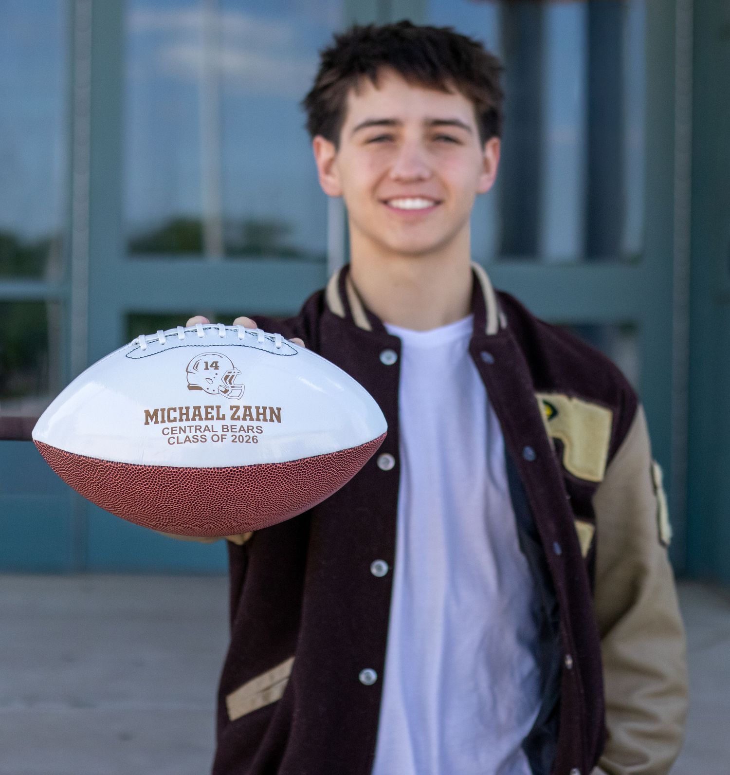 Teenage boy in letterman jacket holding personalized football reading Michael Zahn senior photo.