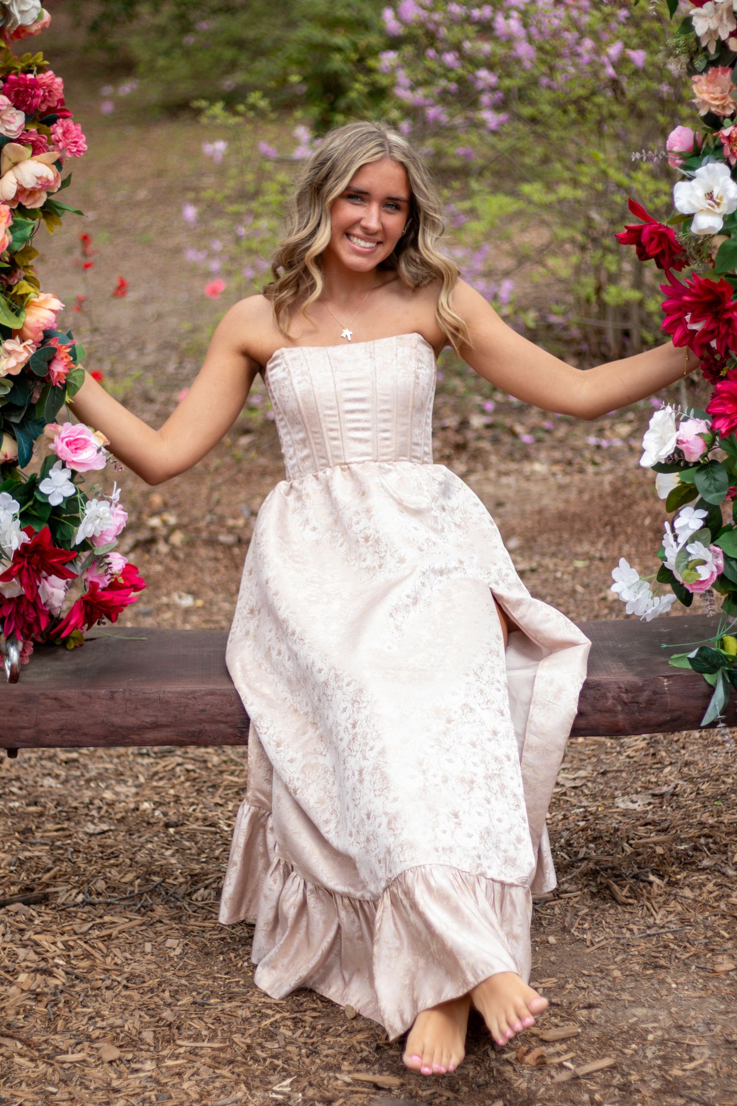 Smiling blonde woman in elegant floral gown sits on a flower-adorned wooden swing in a garden setting.