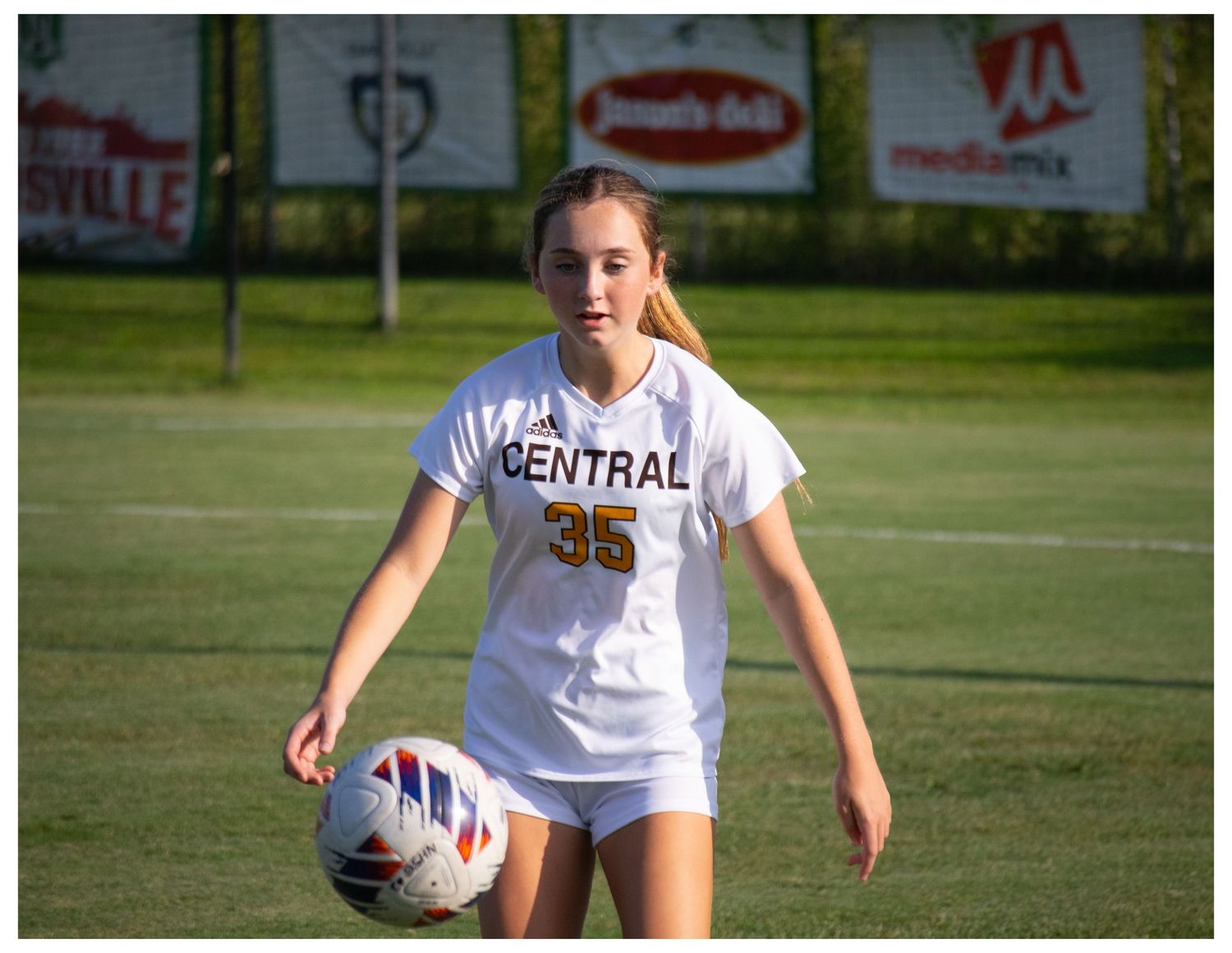 Female soccer player #35 in white Central jersey dribbling ball on green field during outdoor game.