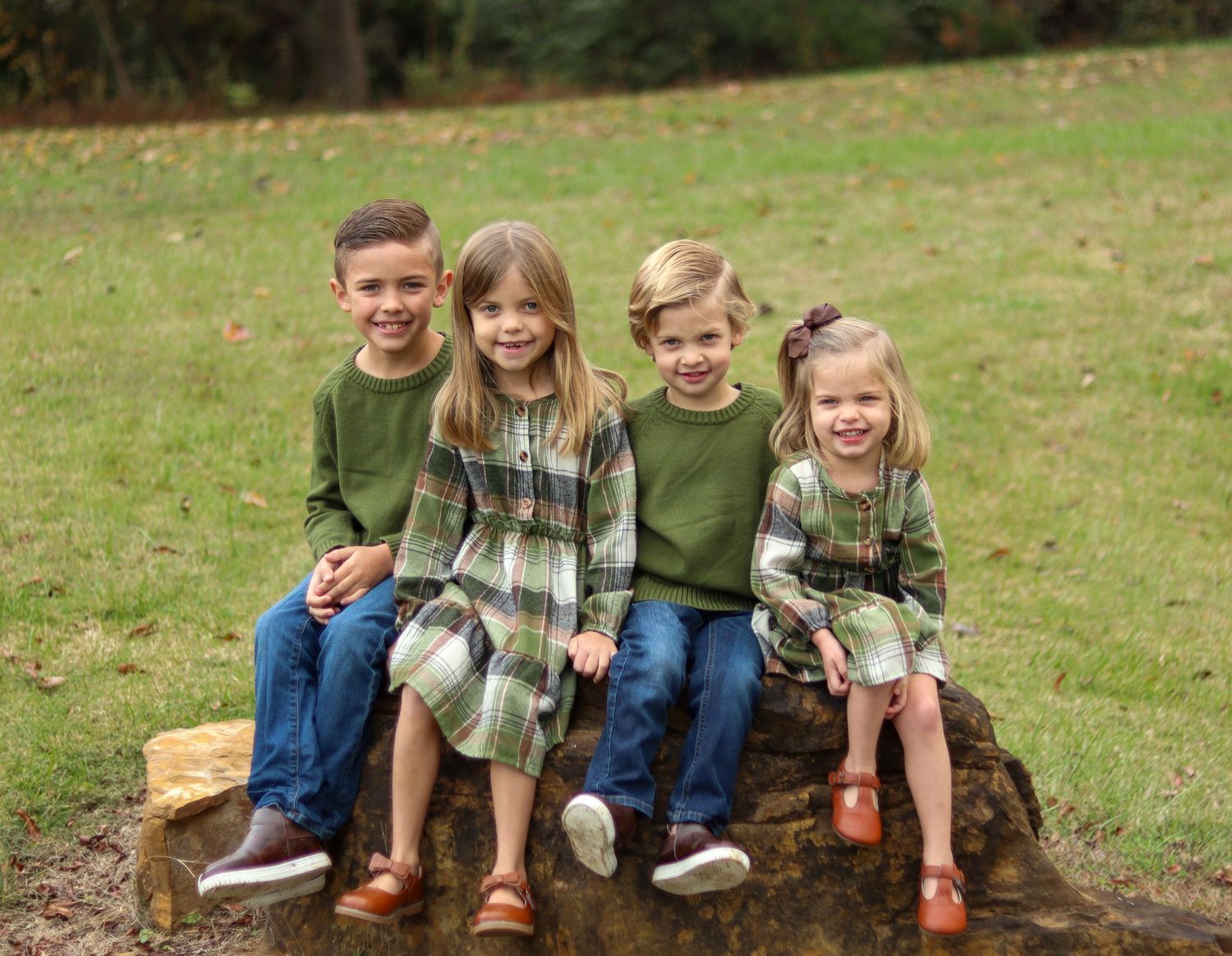 Four happy children in coordinating green outfits sitting together on log in grassy park.