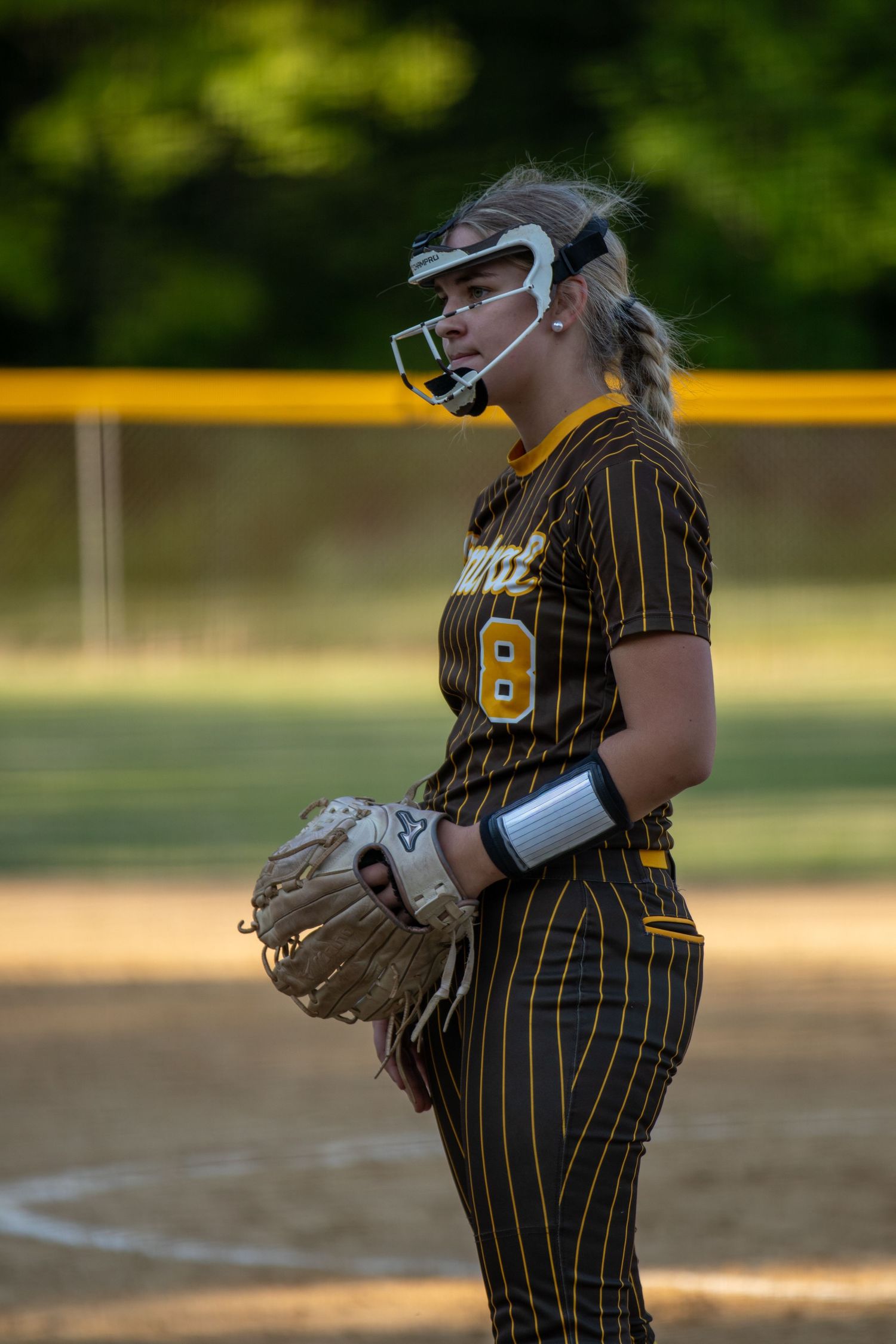 Female softball pitcher #8 in black and gold pinstripe uniform with face guard holds glove on field.