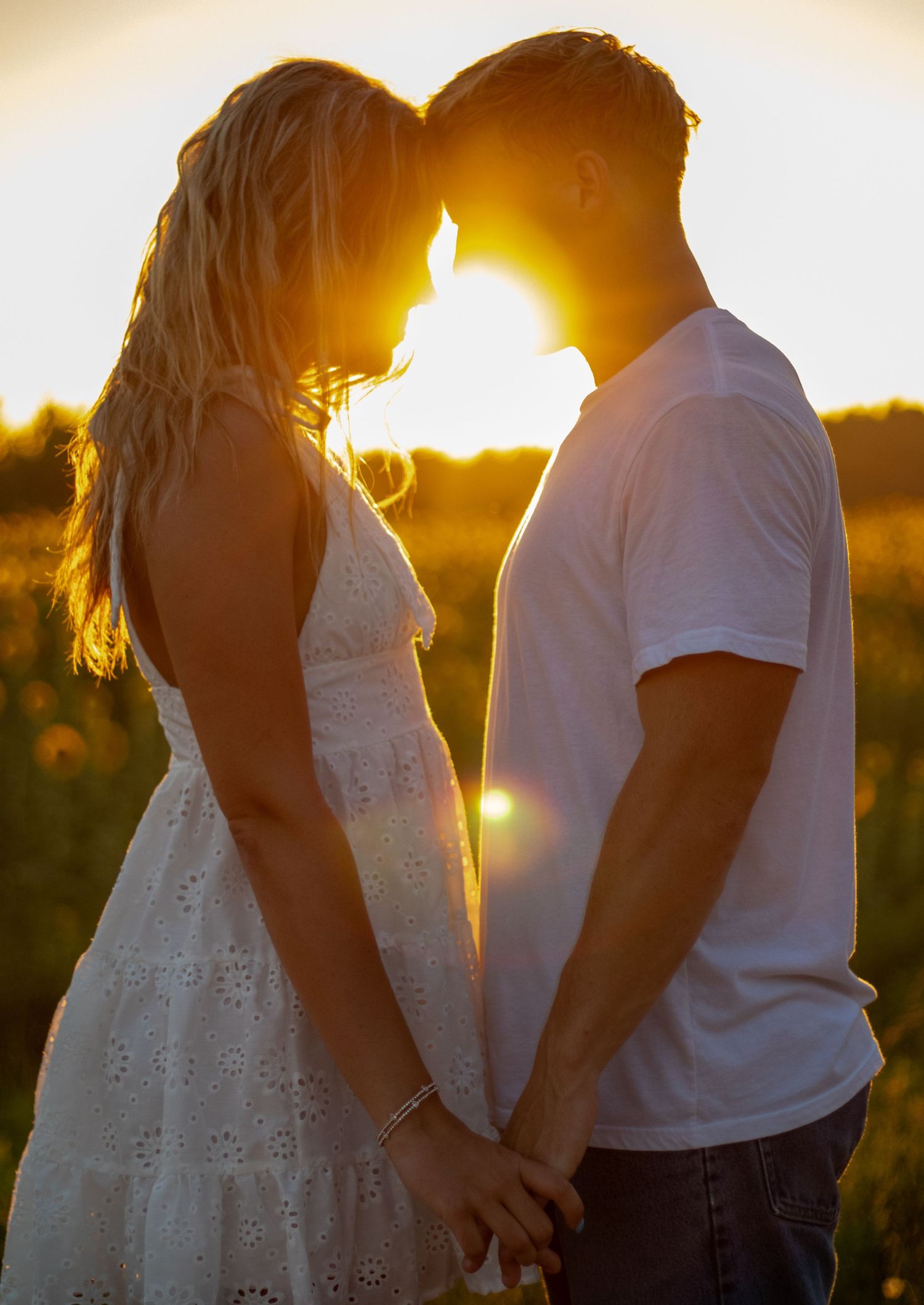 Romantic couple holding hands facing each other with glowing golden sunset behind them in a field.