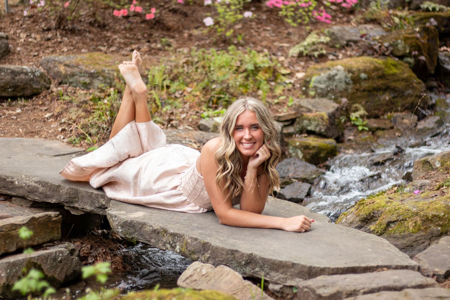 Smiling blonde woman in pink dress poses lying on stone steps beside a scenic waterfall.