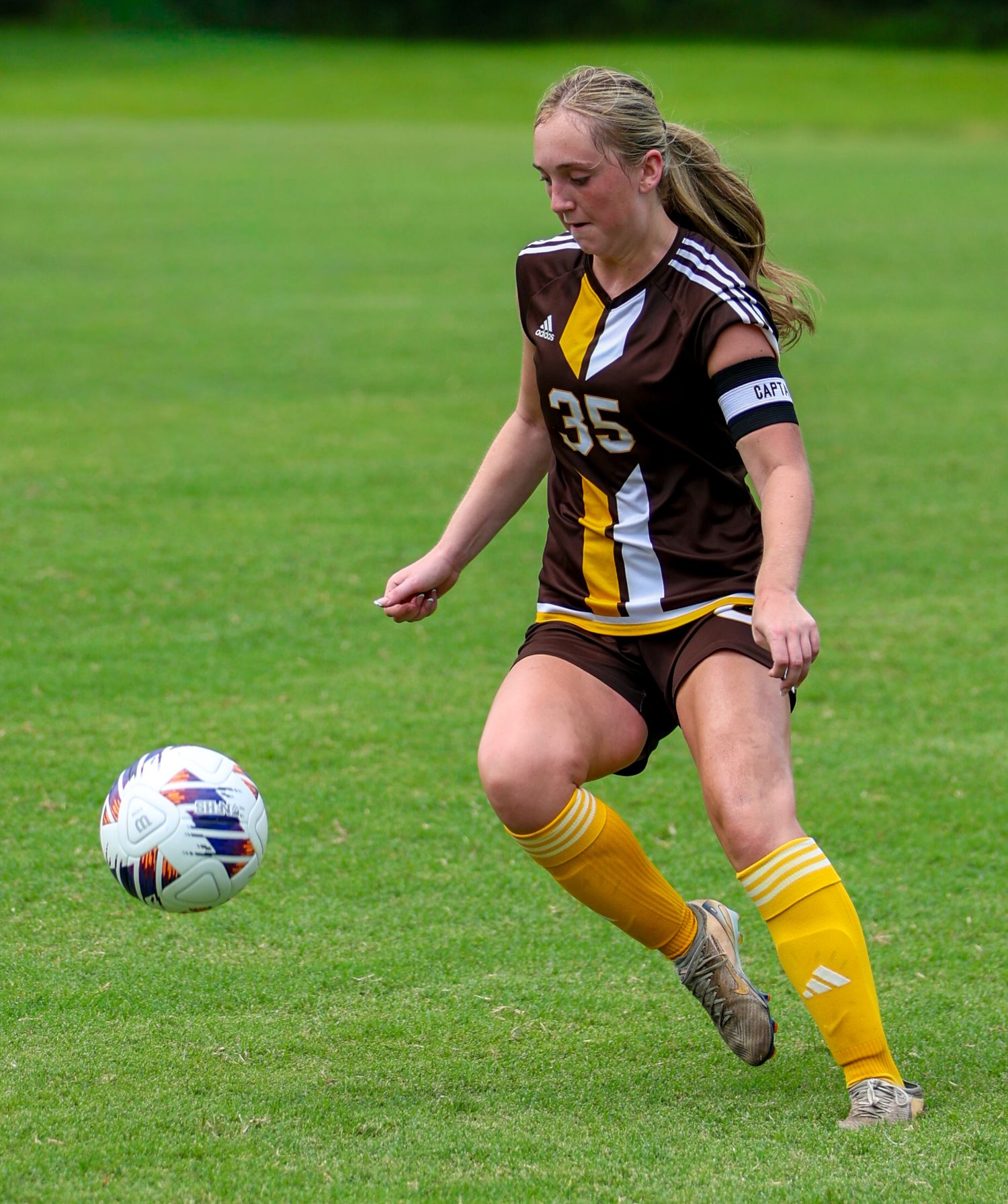 Female soccer player wearing number 35 brown jersey and yellow socks dribbling a ball on a green grass field.