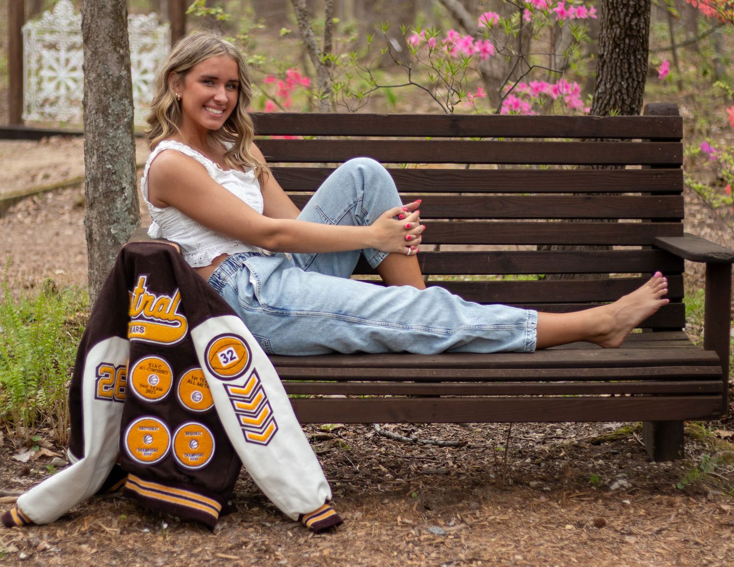 Smiling blonde teen sitting on bench with letterman jacket in spring garden setting.
