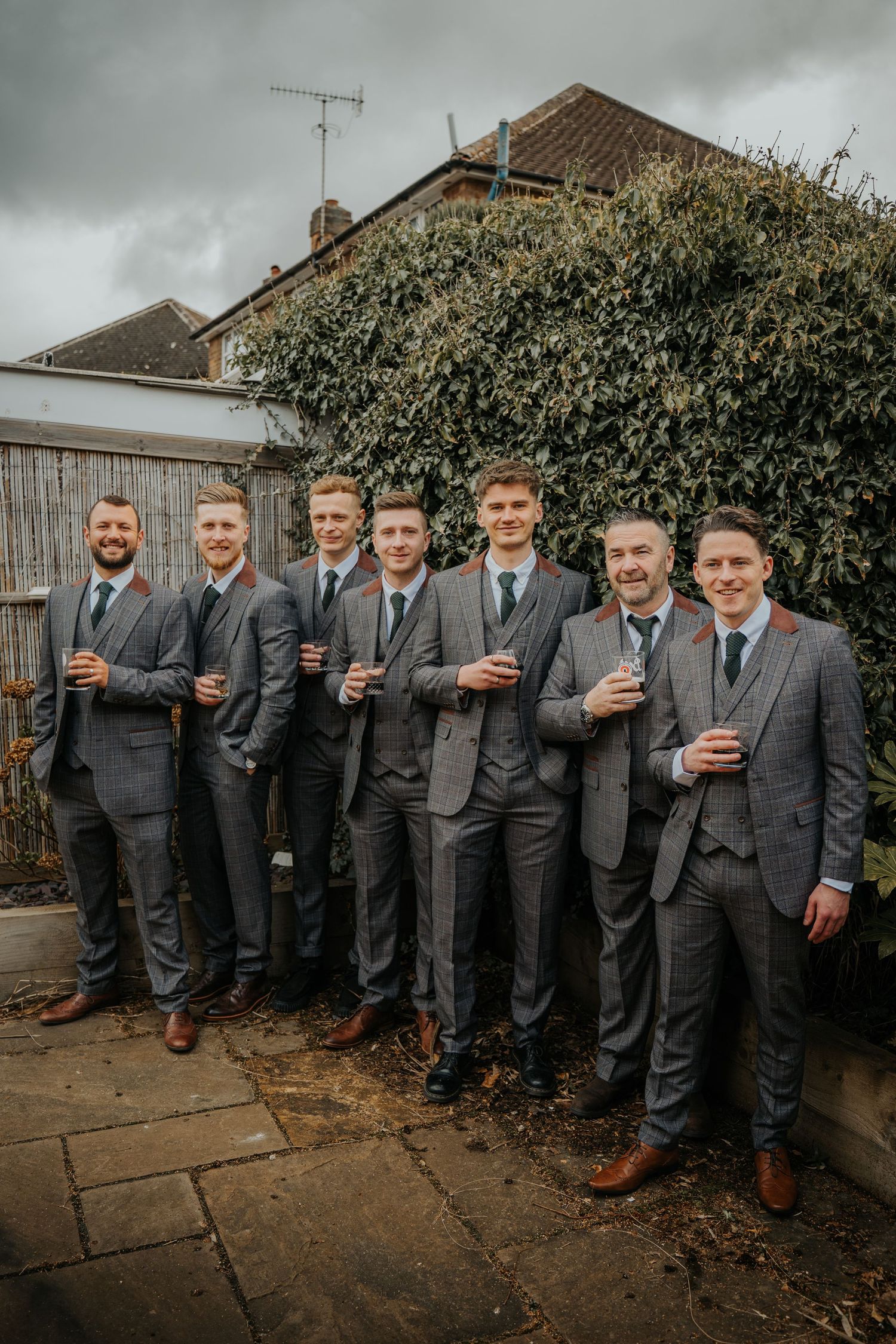 Seven groomsmen in matching grey suits with red boutonnieres pose outdoors holding drinks at a wedding.