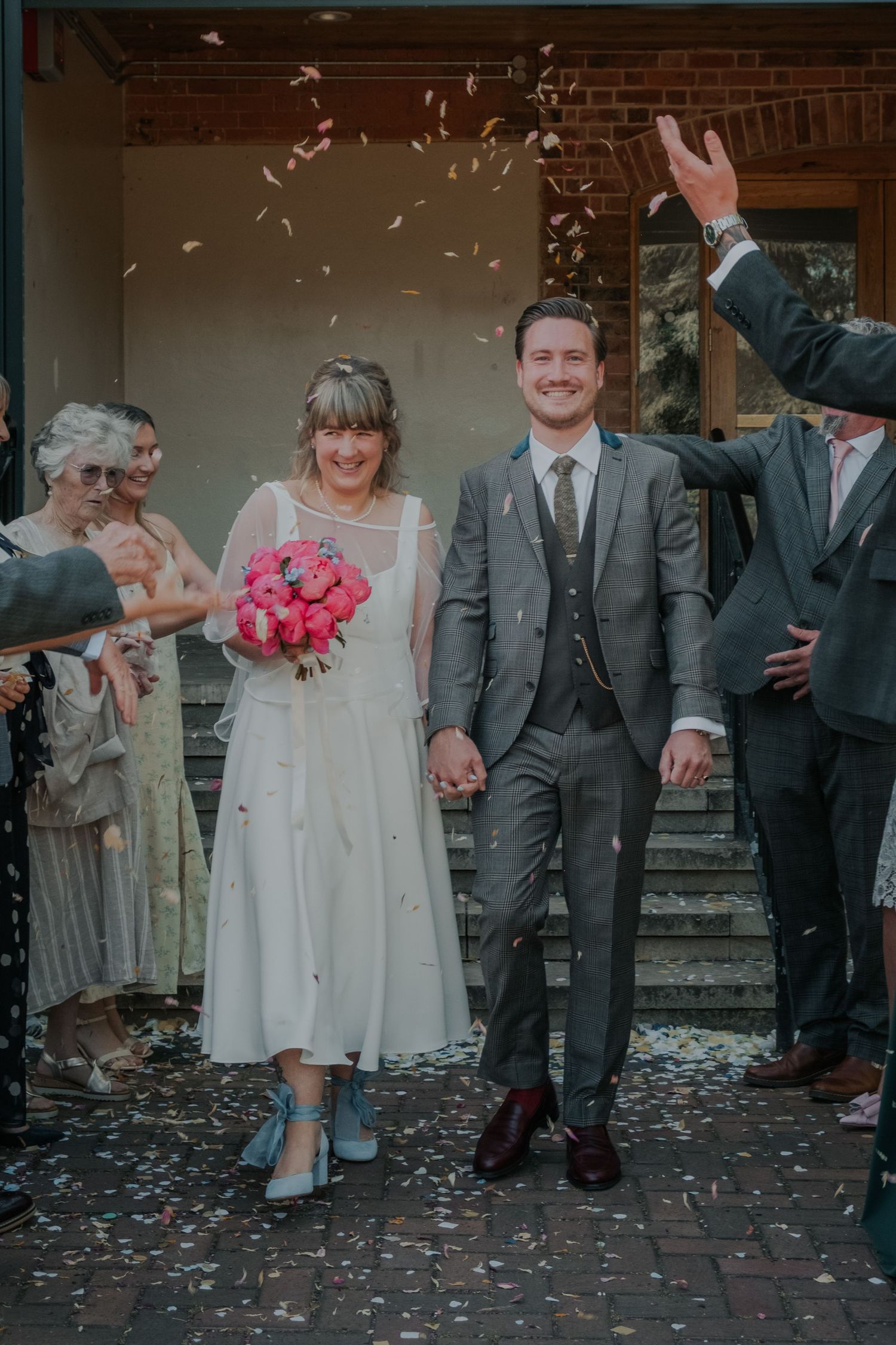 Joyful newlyweds walking through confetti shower outside ceremony venue holding pink bouquet.
