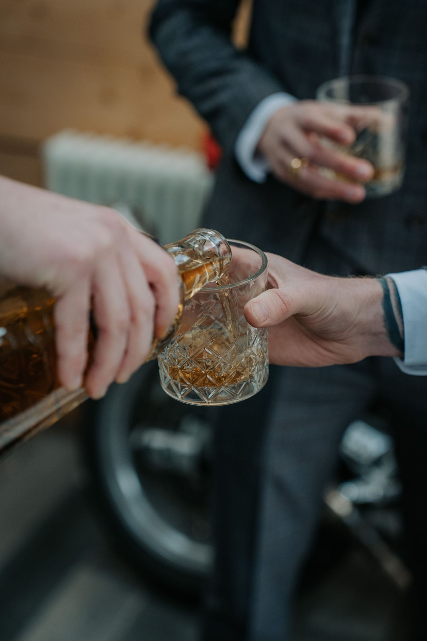Close-up of hands pouring amber whiskey from a crystal decanter into a rocks glass at a formal gathering.