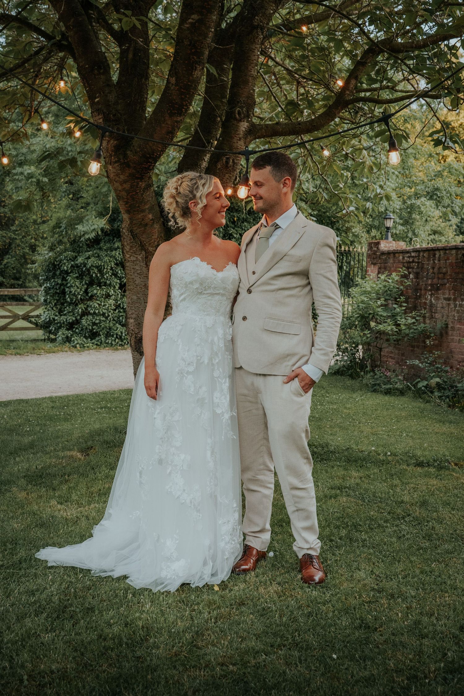 Bride in white lace gown and groom in beige suit stand together on green lawn under fairy lights.