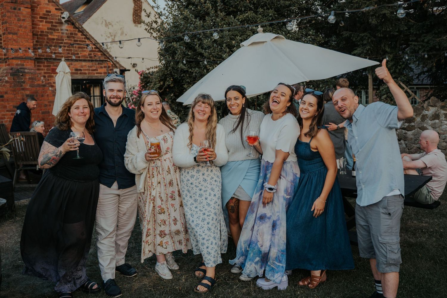 Group of friends smiling outdoors at a garden party, holding drinks near a brick building on a sunny day.