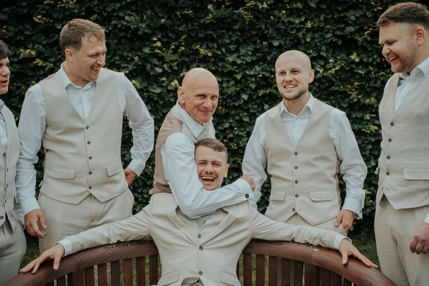 Groom and groomsmen laughing together in matching beige suits and vests at an outdoor wedding celebration.