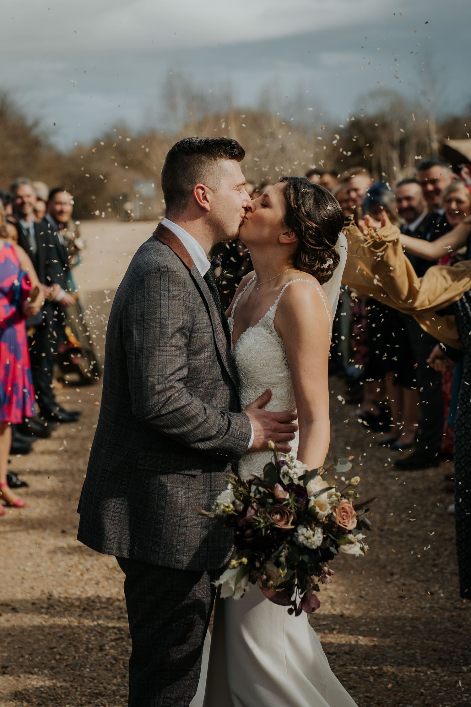 Bride and groom share a kiss during confetti shower, surrounded by wedding guests outdoors on their special day.