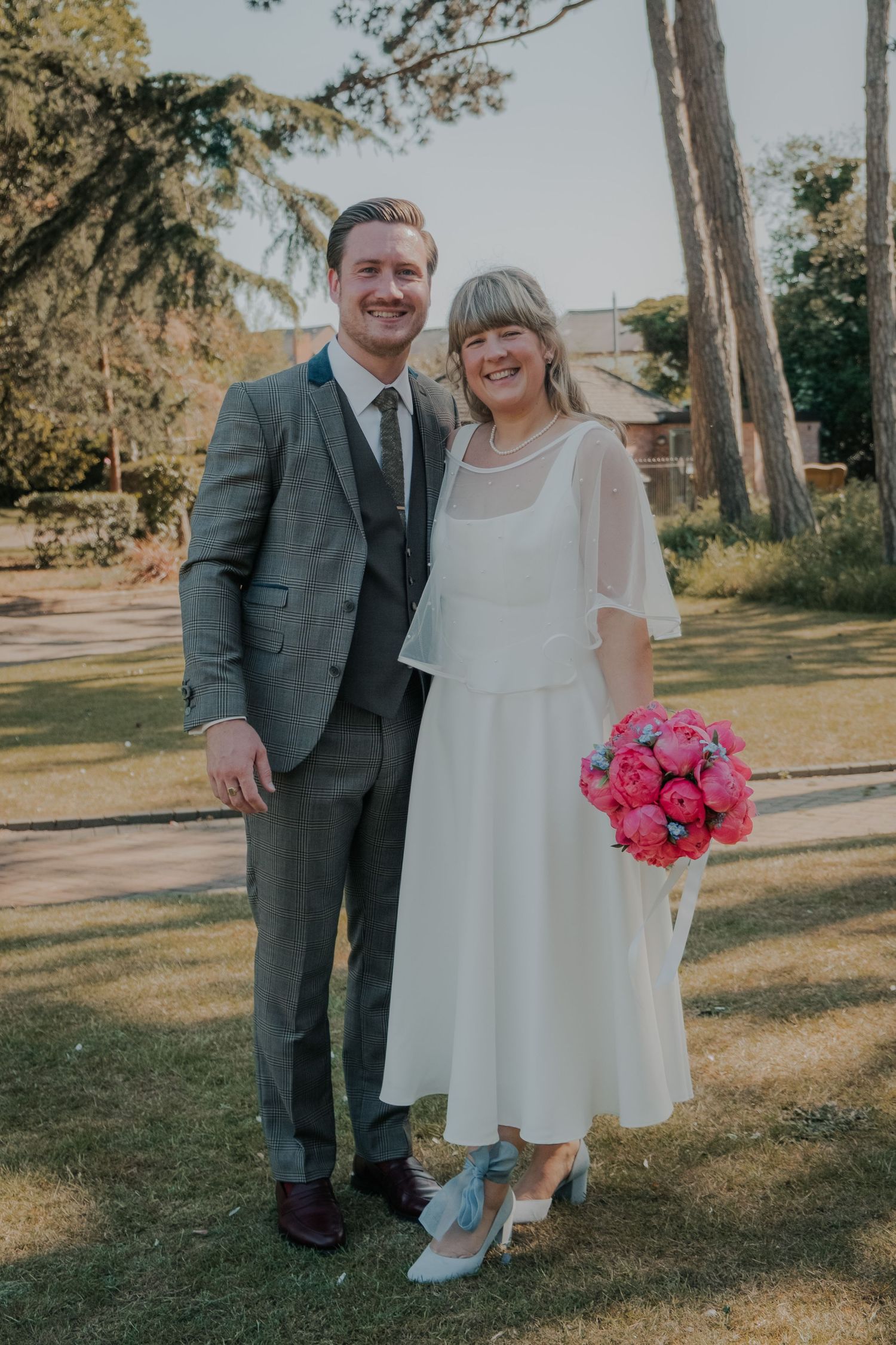 A smiling bride in white midi dress holding pink bouquet stands with groom in gray suit outdoors on green lawn.