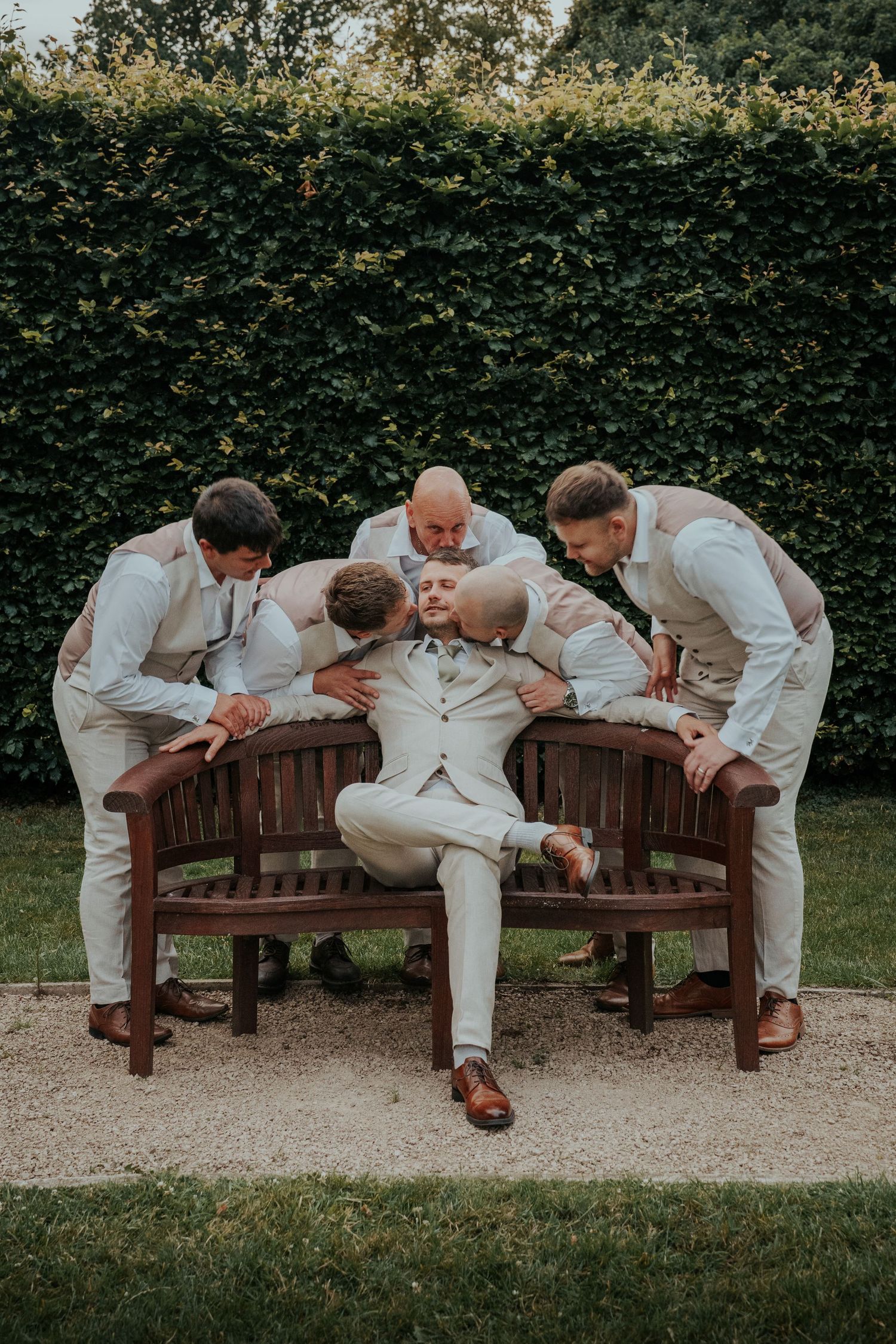 Groom in white suit relaxes on wooden bench surrounded by groomsmen in matching attire, posing outdoors near green hedge.