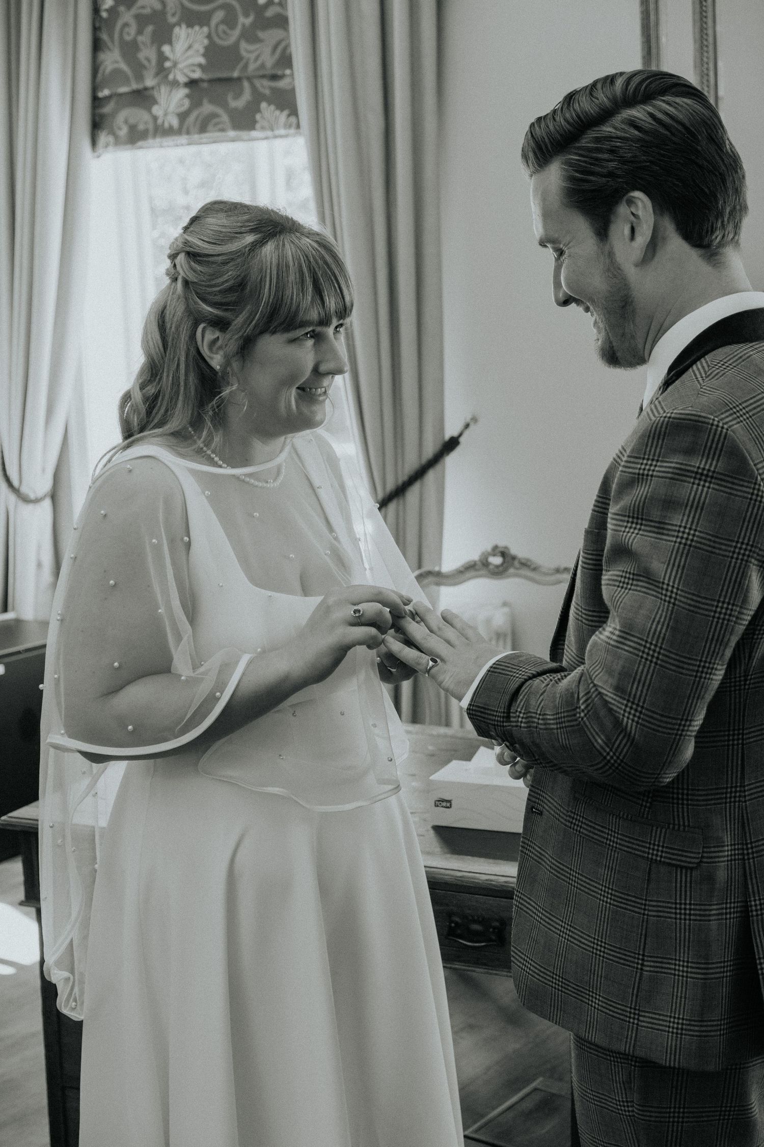 Black and white photo of a bride and groom exchanging rings during their wedding ceremony indoors.