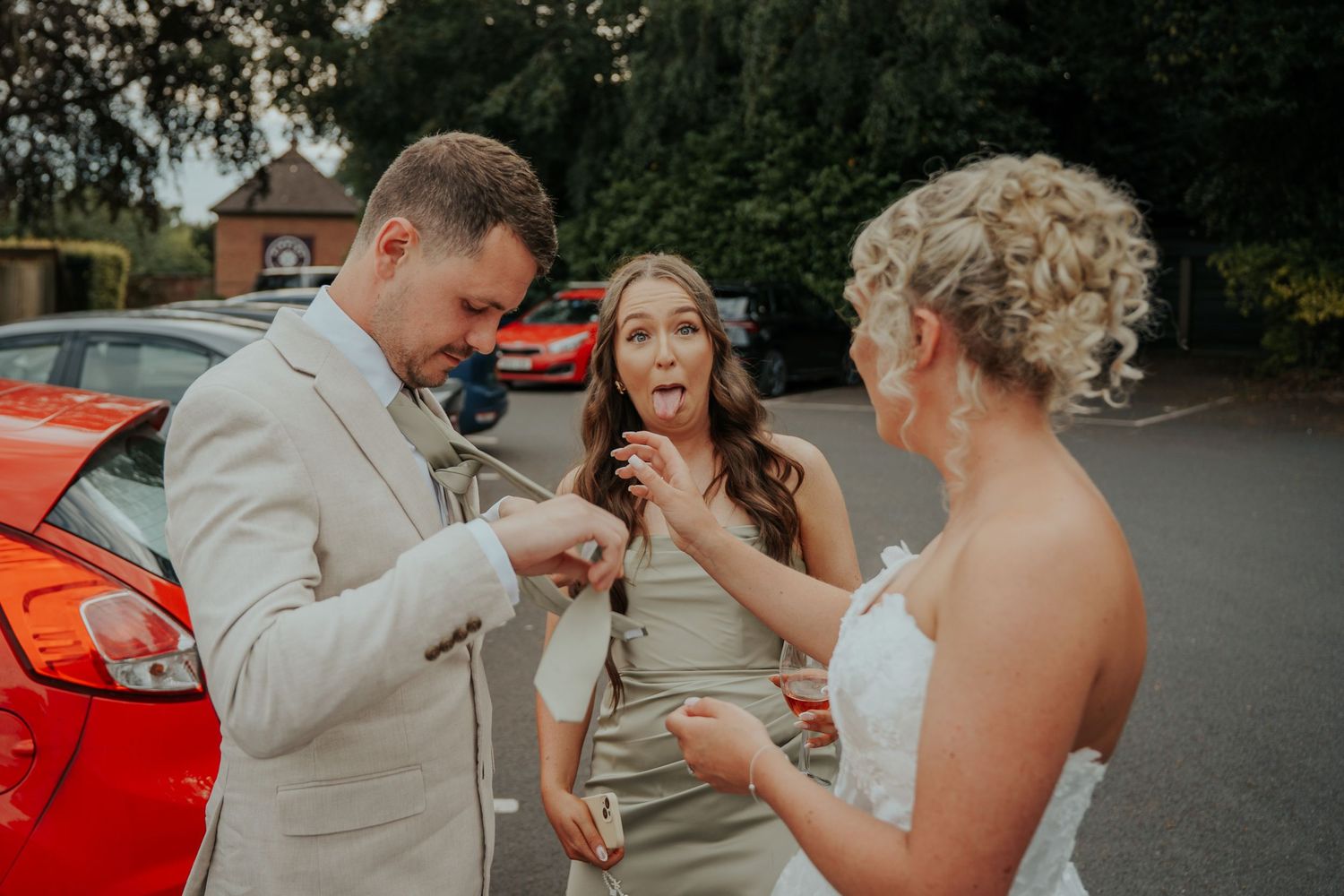 Bride and groom with bridesmaid laughing beside a red car at a wedding venue outdoor setting.