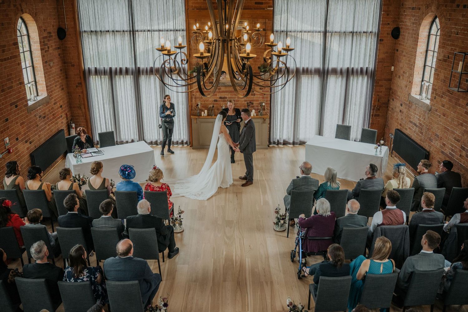 Elegant wedding ceremony in brick venue with chandelier, couple kissing at altar, guests seated in rows.
