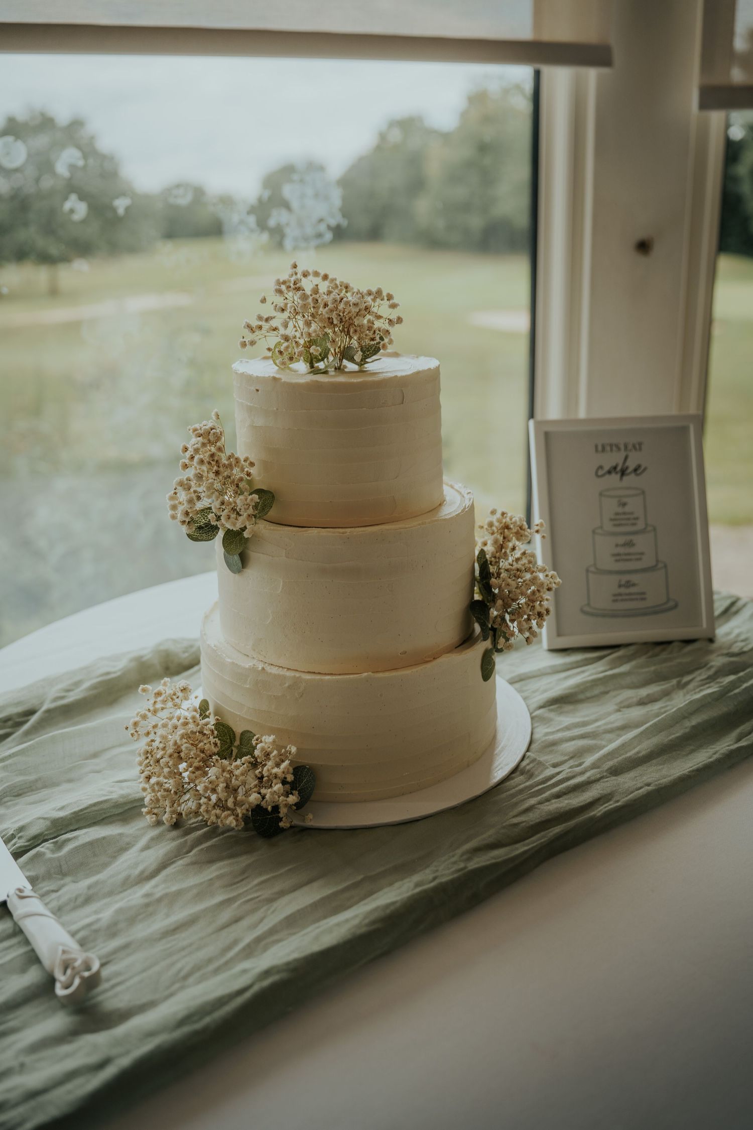 Three-tier white wedding cake with baby's breath and greenery florals on green linen table runner by window.
