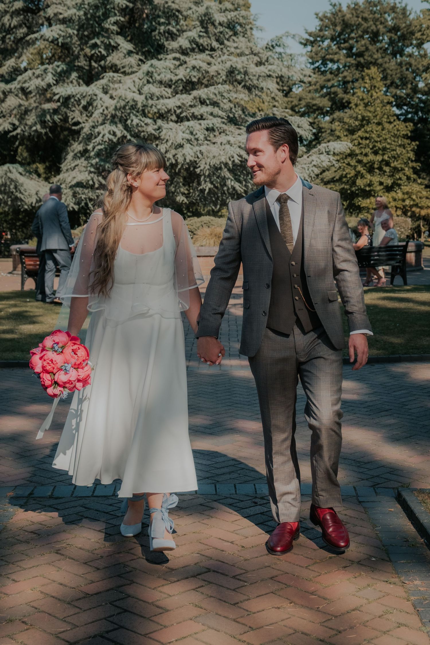 Bride and groom walking hand in hand in a park, she holds pink bouquet, he wears a grey suit with burgundy shoes.