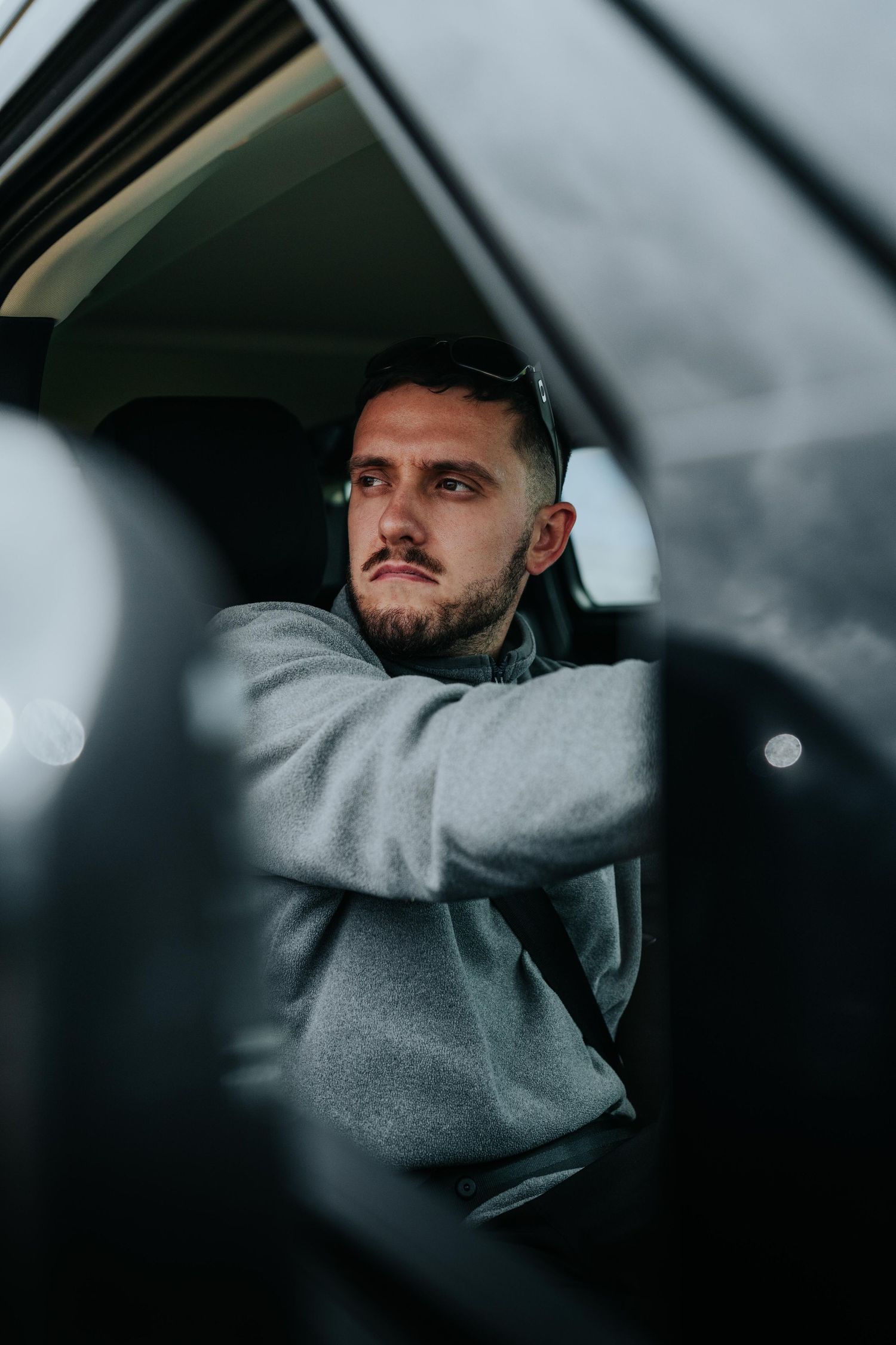 A young bearded man in a gray hoodie sits in a car, arms crossed over the steering wheel, looking thoughtfully ahead.