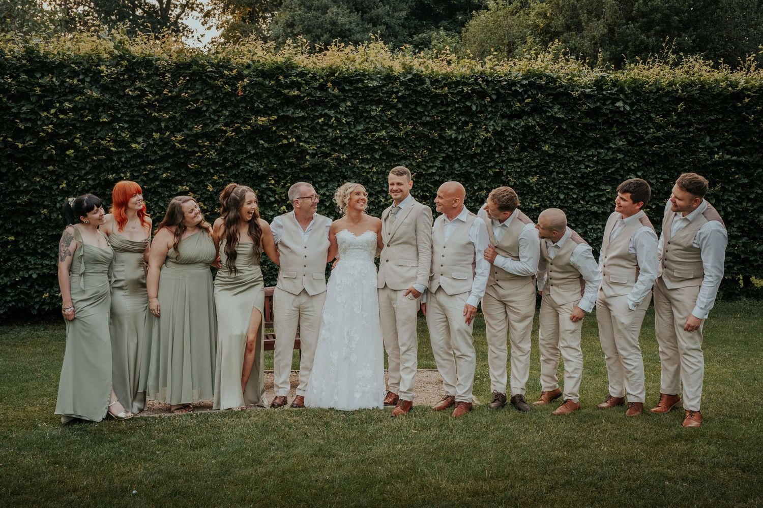 Wedding party in sage green and beige attire posing together on lawn with hedge backdrop.