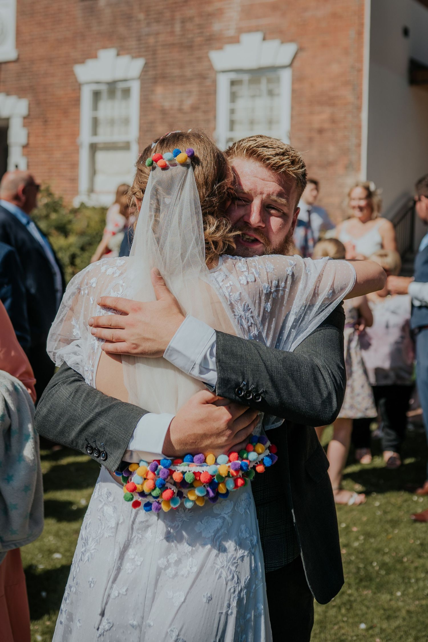 Groom hugging bride in colorful pom-pom trimmed wedding dress and veil outside brick church.