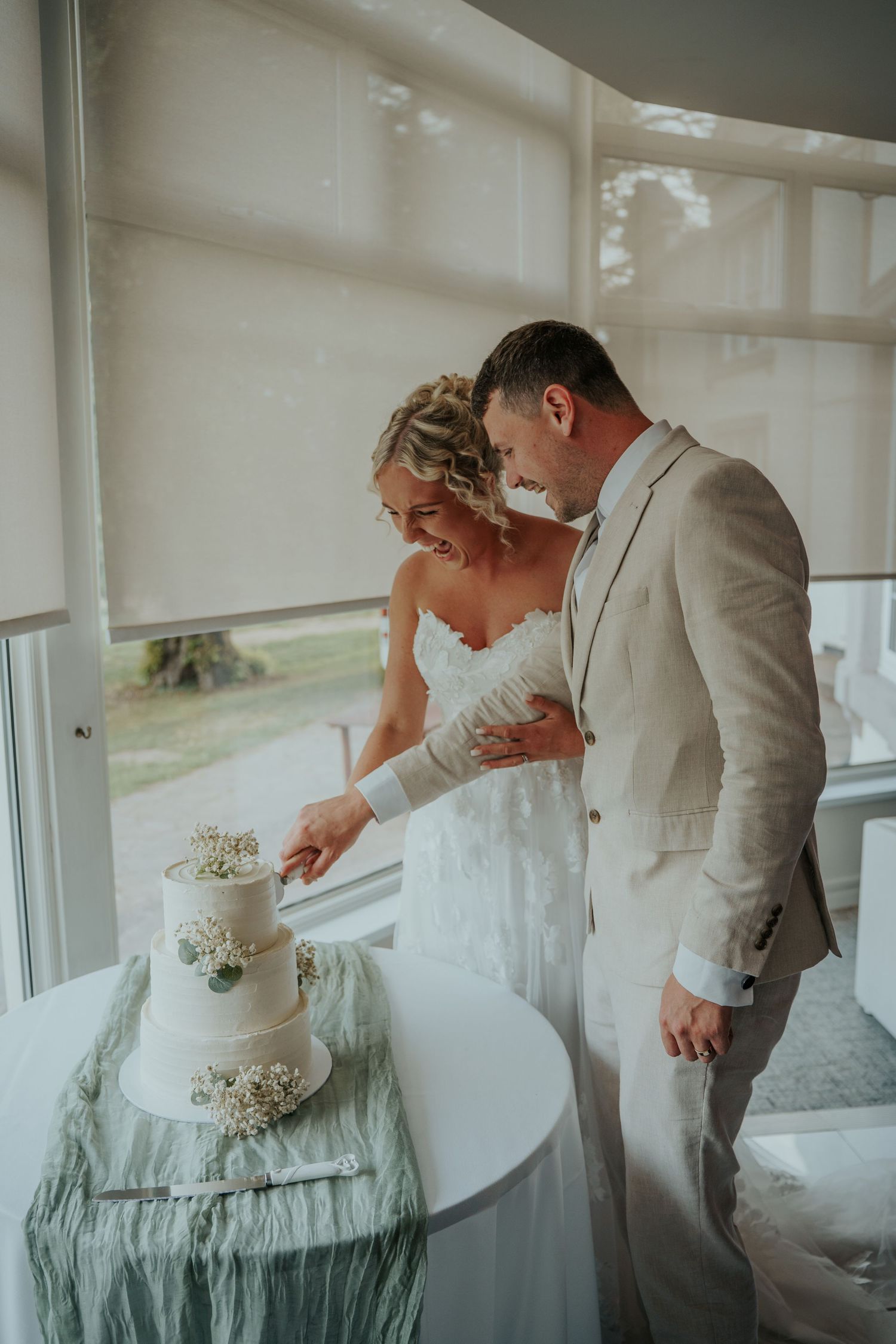 Bride and groom cutting their white three-tier wedding cake together, smiling, in a bright venue with large windows.
