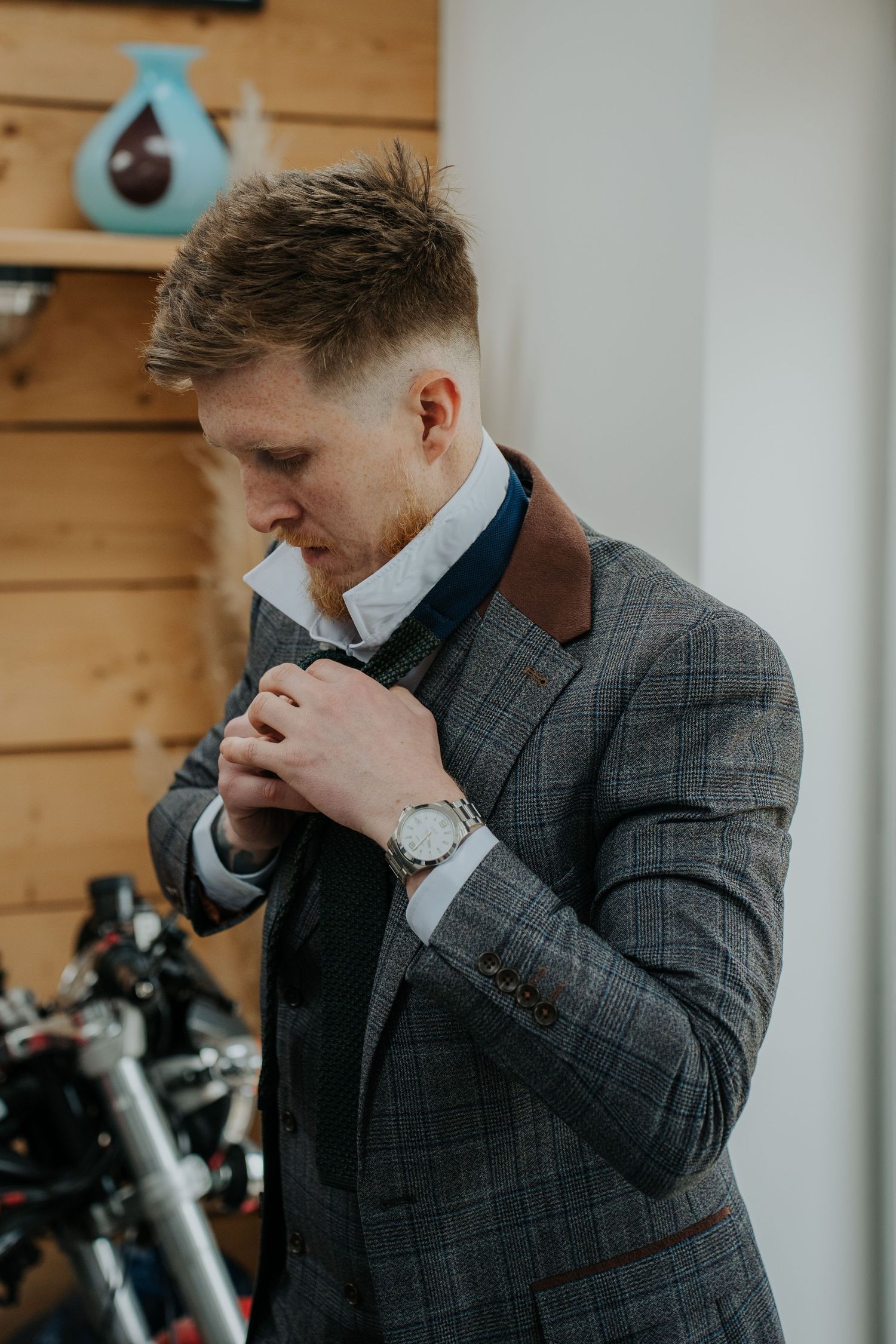 Man in gray plaid suit adjusting tie, wearing silver watch, standing indoors near wooden wall.