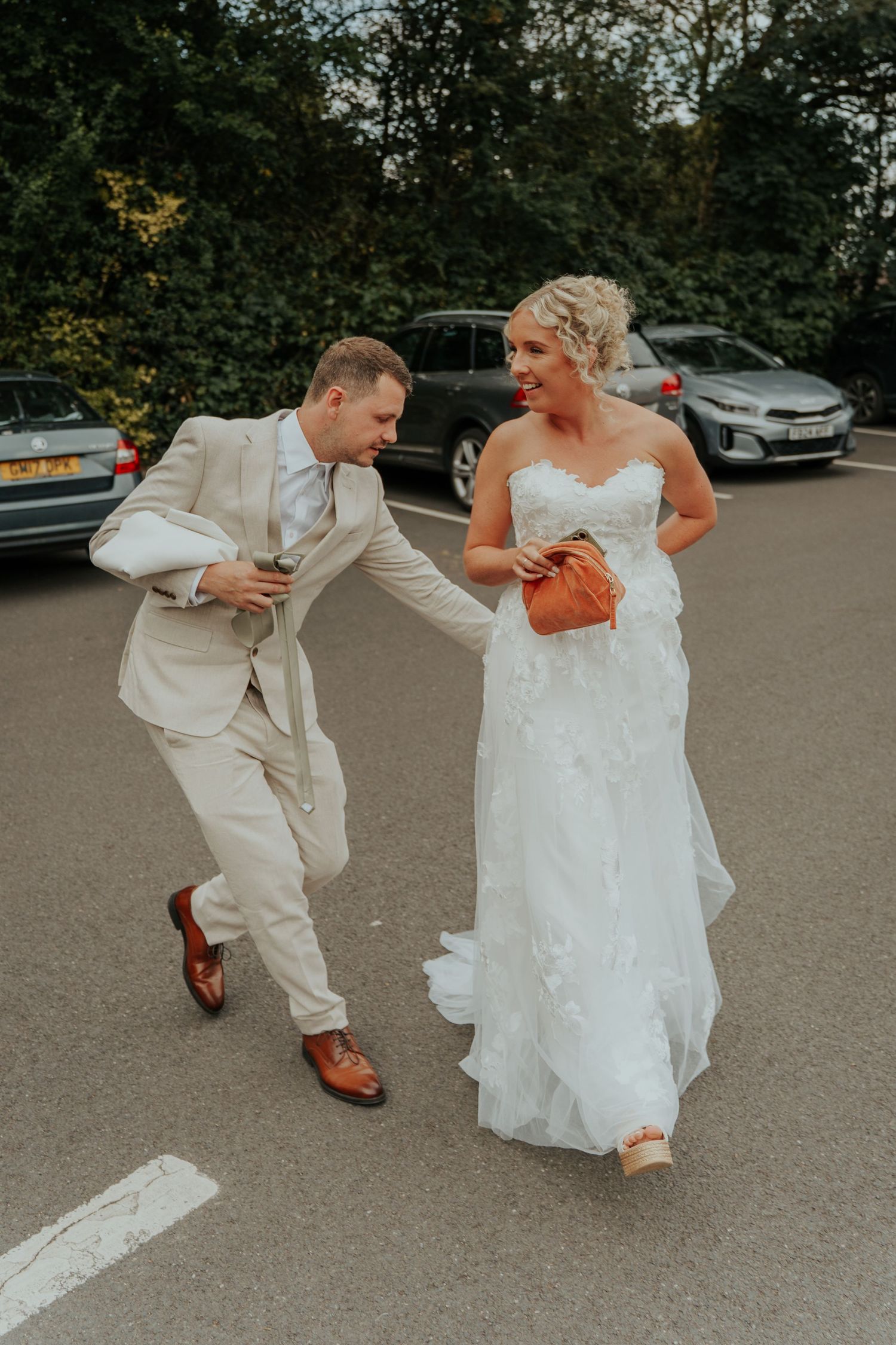 Laughing bride and groom stroll through a parking lot, sharing a playful moment on their wedding day.
