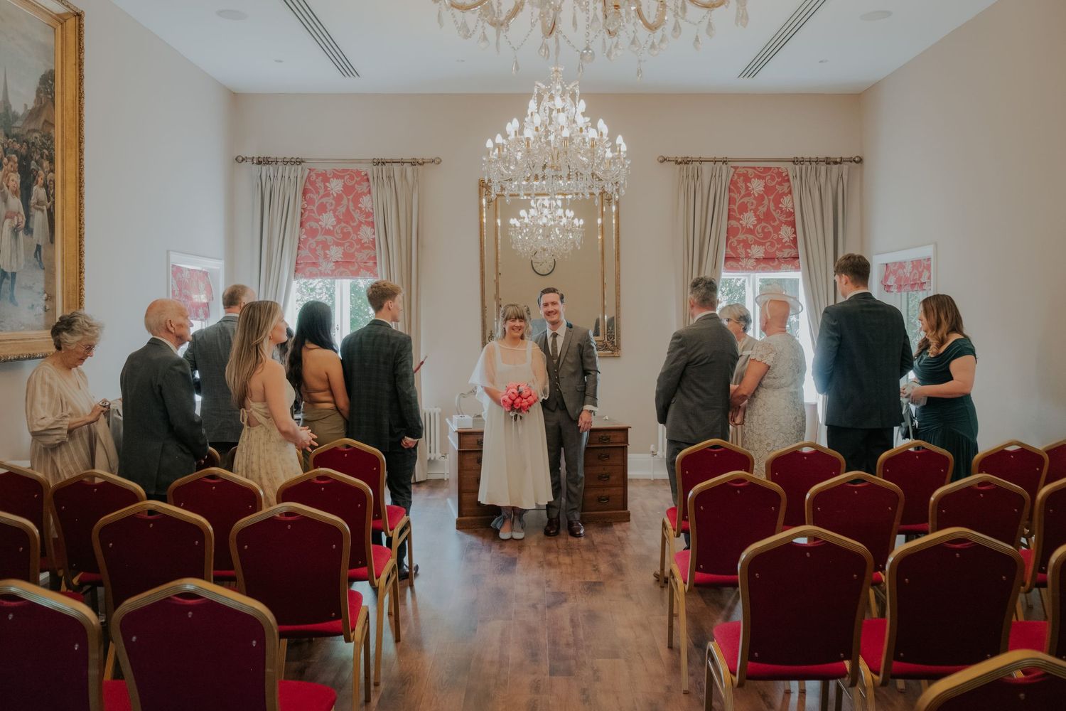 Elegant wedding ceremony in a grand room with red chairs, chandelier, and couple at the altar.
