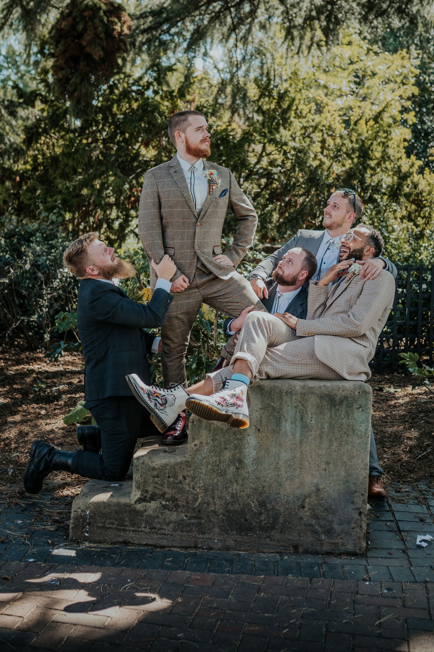 Groom in plaid suit poses with groomsmen on stone steps outdoors, one kneeling, others seated, all in wedding attire.