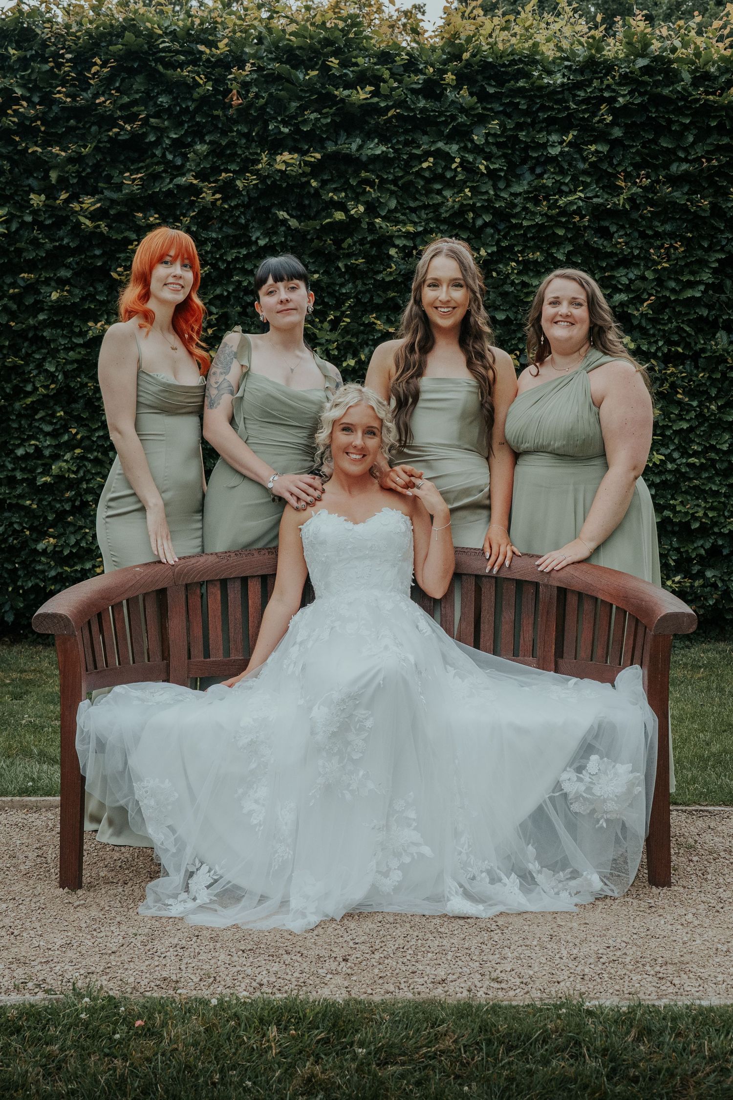 Bride in white gown sits on wooden bench surrounded by four bridesmaids in sage green dresses outdoors.