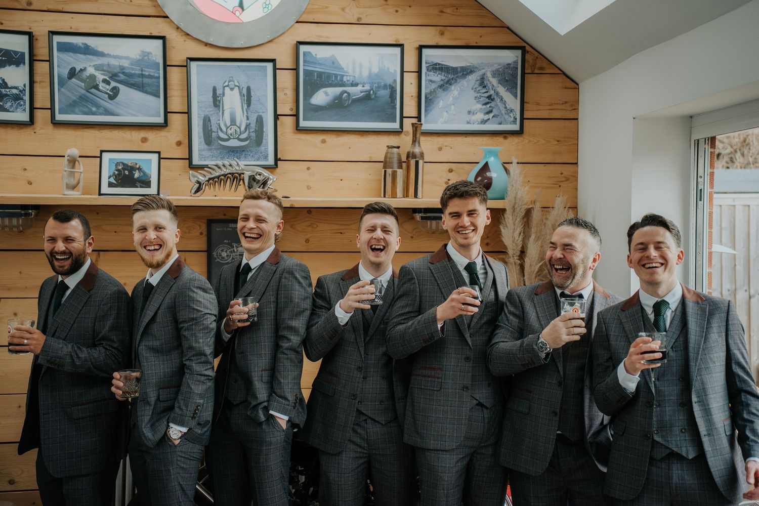 Seven groomsmen in matching grey suits laughing and raising glasses in a rustic wood-paneled room with vintage racing artwork.