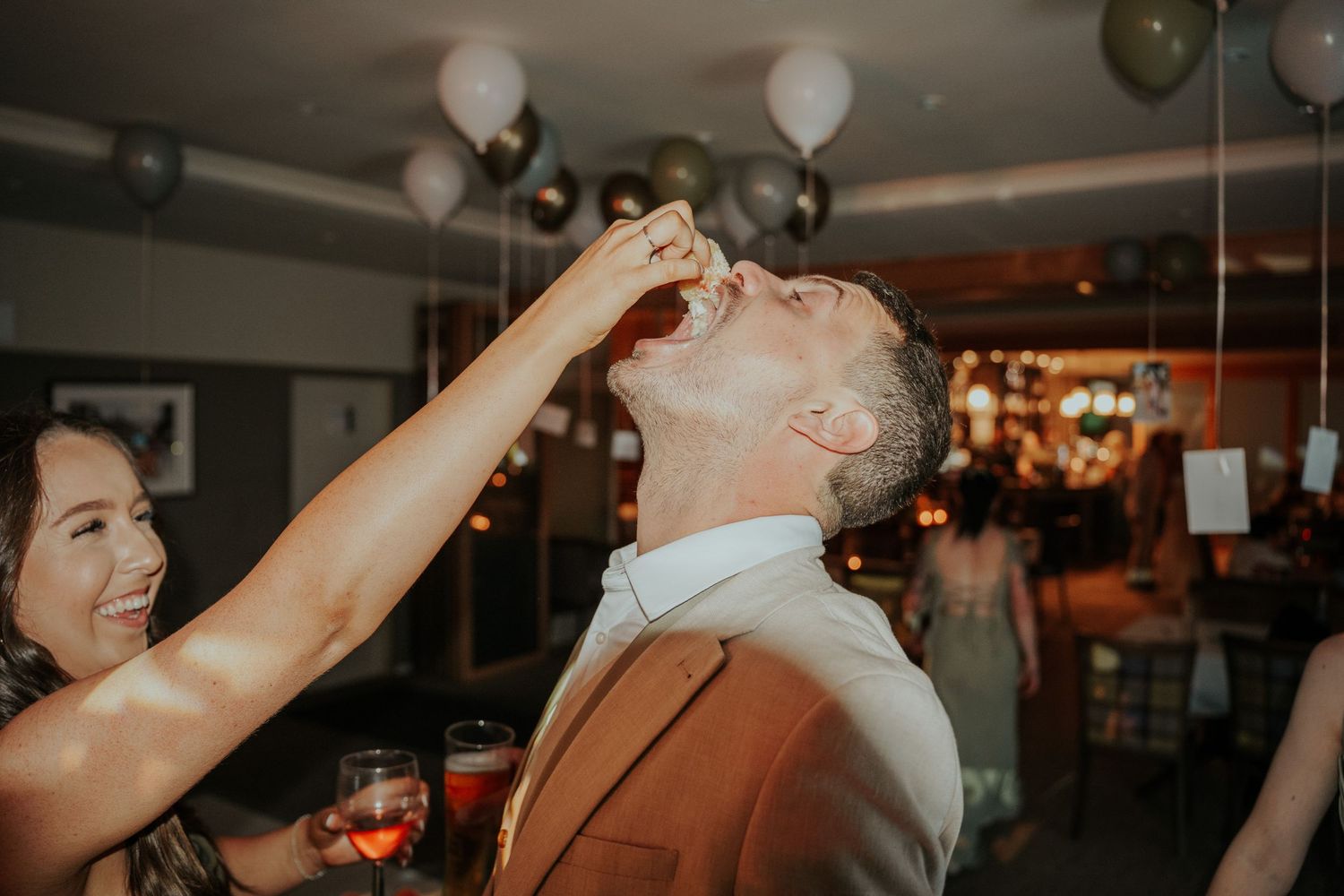 Woman feeding a shot to a man in a brown suit at a festive party with balloons in the background.