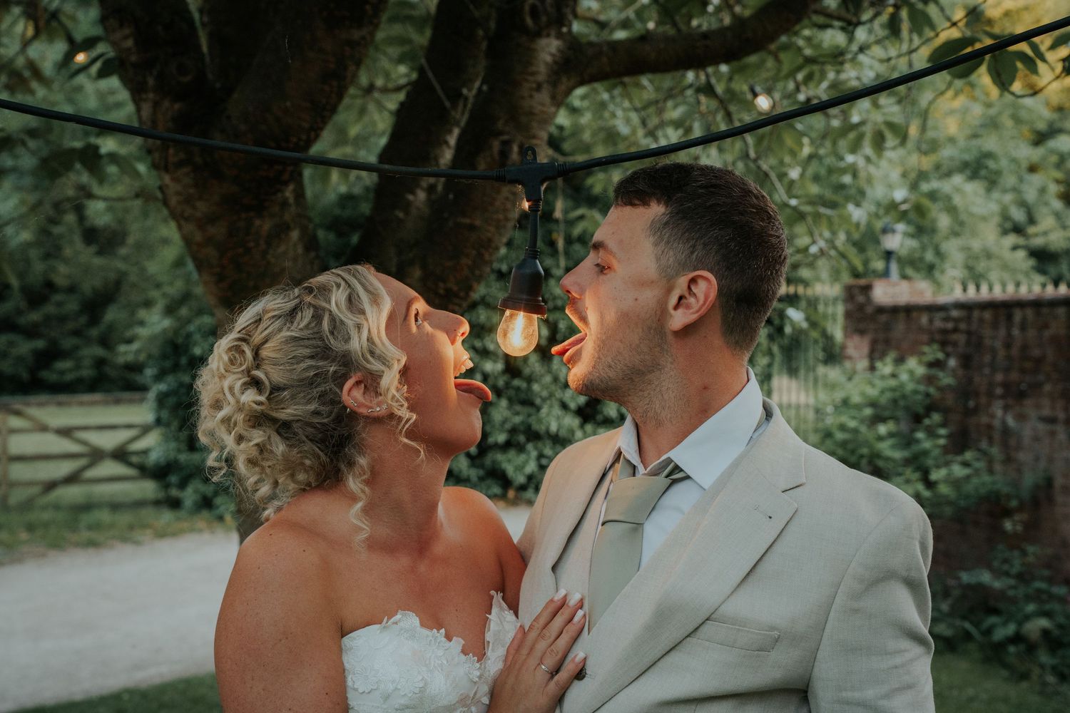 Bride and groom sharing playful romantic moment under string lights in garden at golden hour.