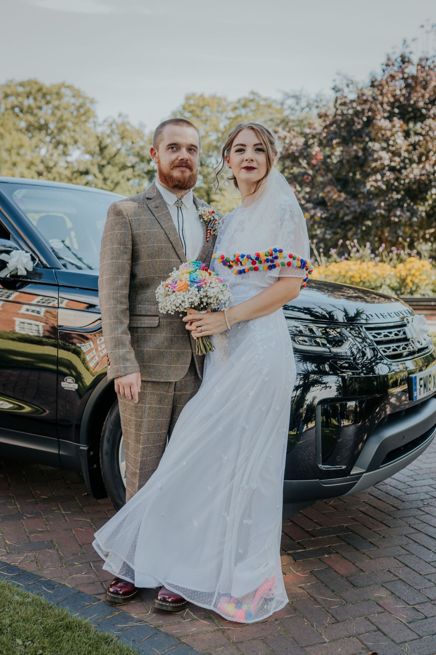 Bride in white gown with colorful details and groom in tweed suit pose by a black Land Rover on their wedding day.
