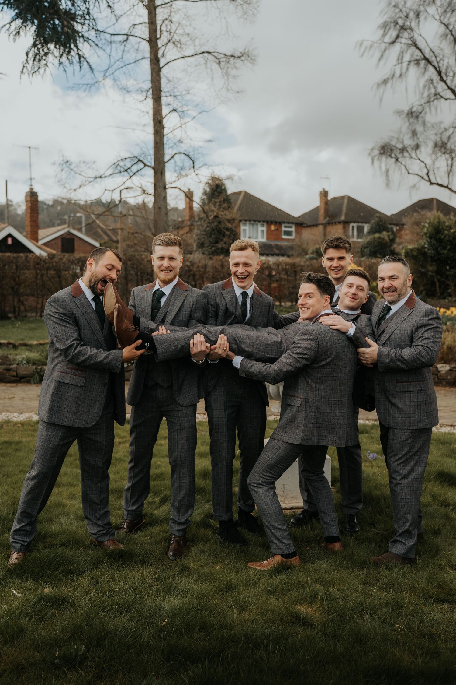A group of groomsmen in grey suits laugh and pose together outdoors at a wedding celebration.