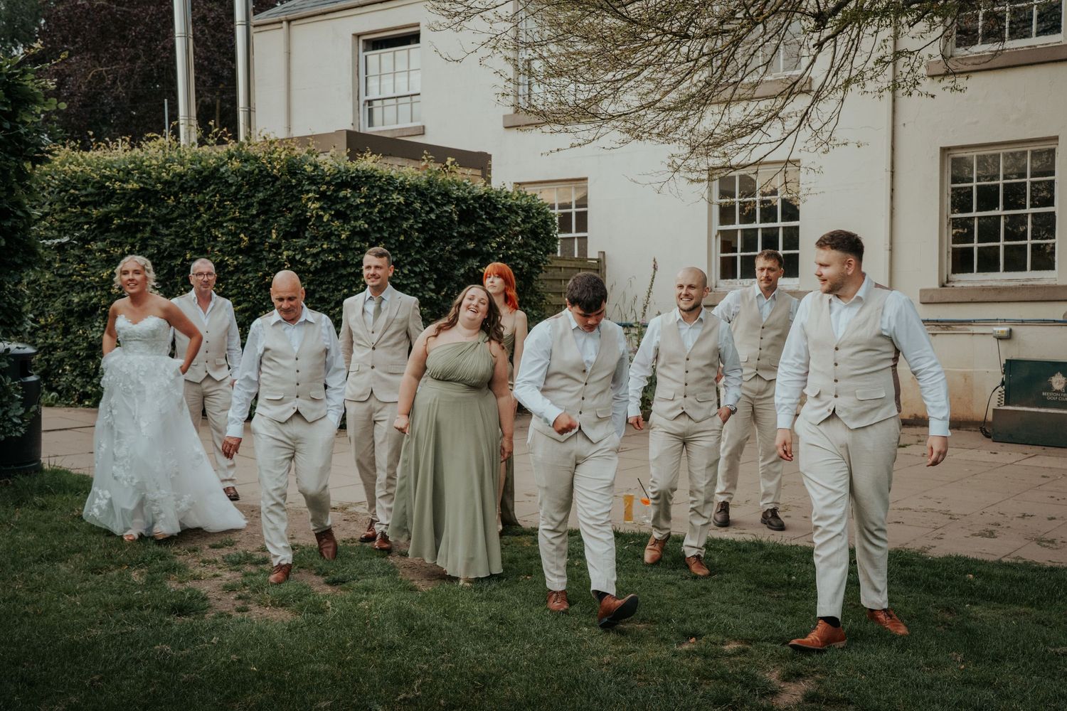 Wedding party walks outdoors in coordinated sage green and neutral attire beside a classic white manor house.