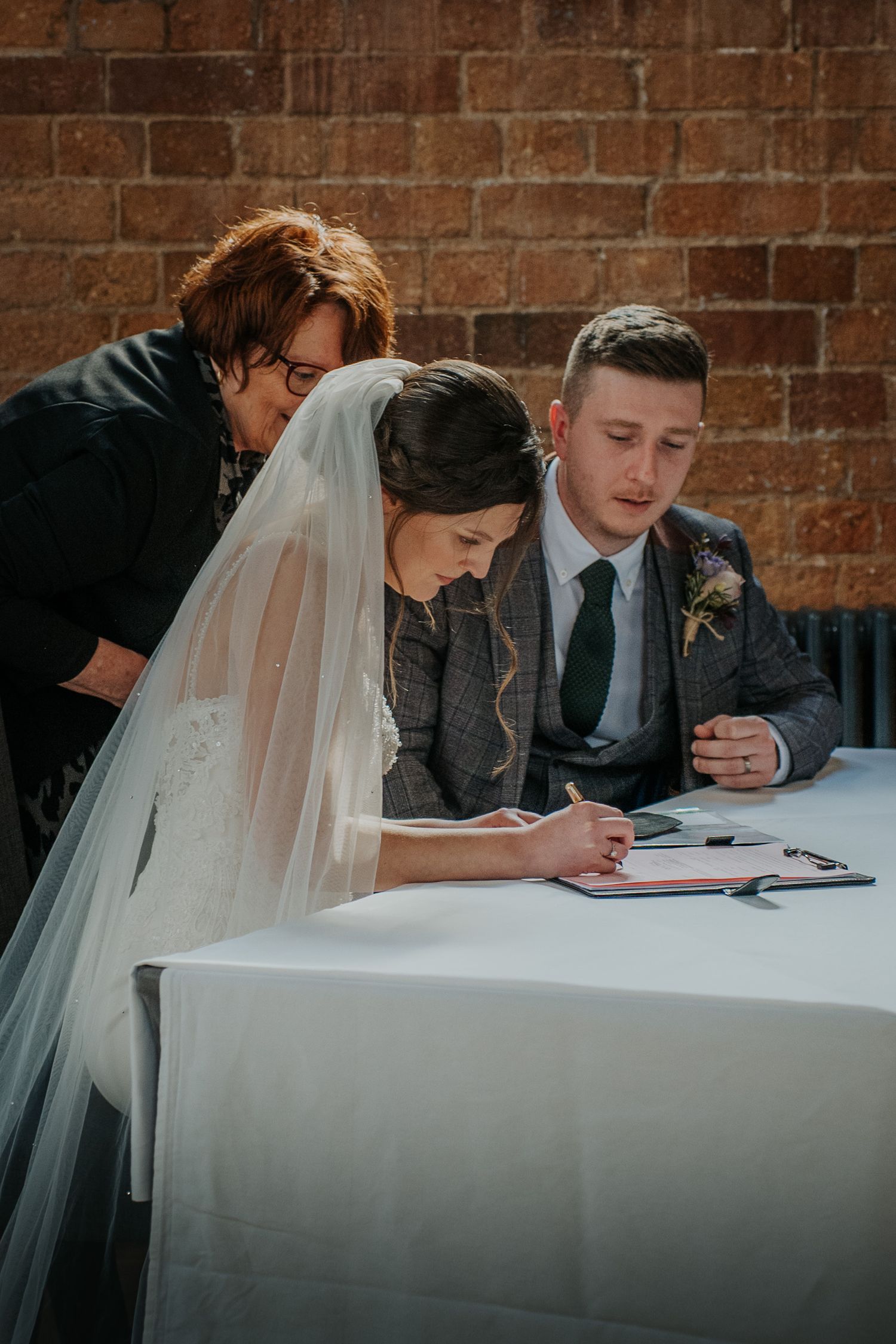 Bride signing wedding register at table with groom and officiant, wearing veil, in front of brick wall.