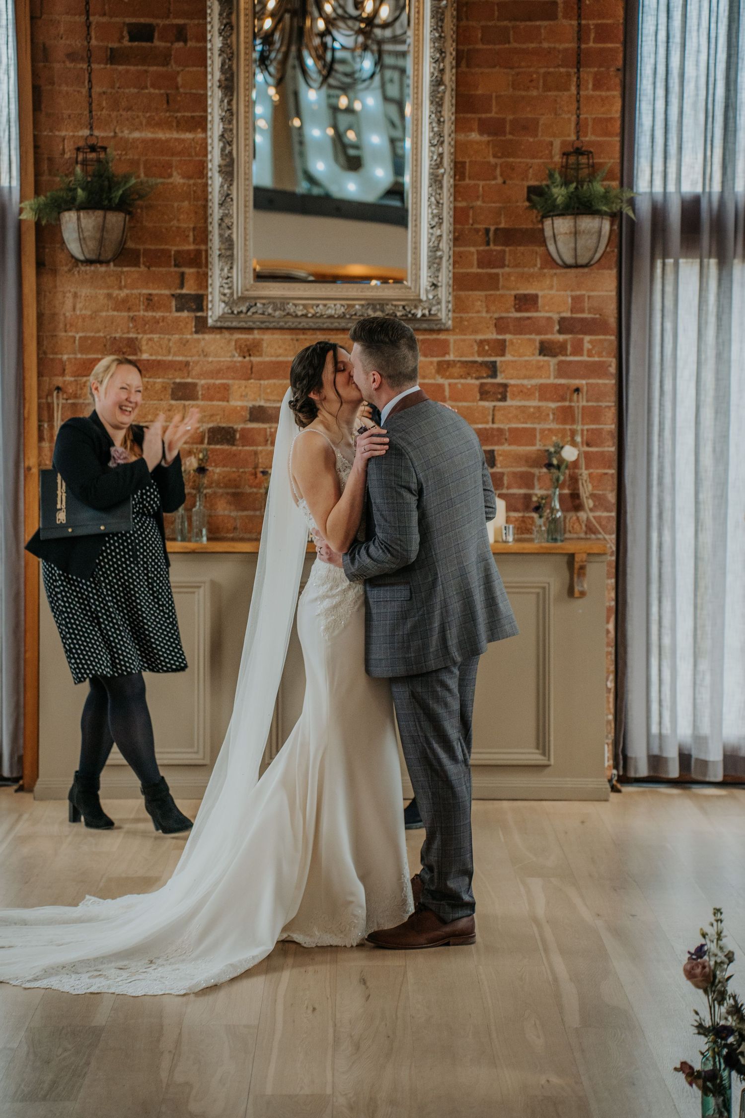 Bride and groom share first kiss at rustic brick wall venue, with celebrant clapping in background.