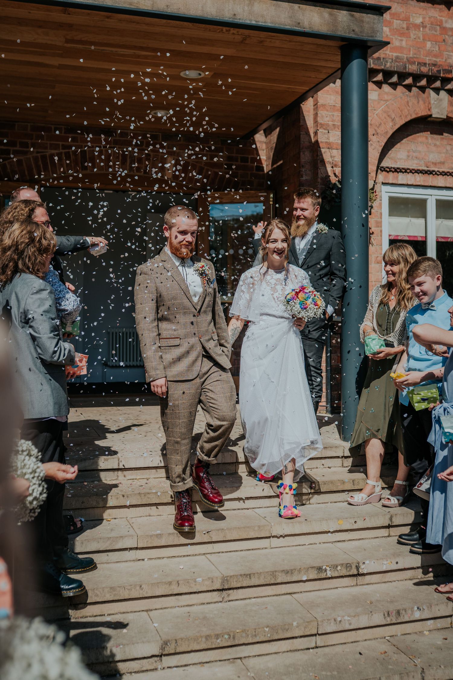 Bride and groom walk through confetti shower outside brick venue, guests cheering, bride in white dress, groom in tan tweed suit.